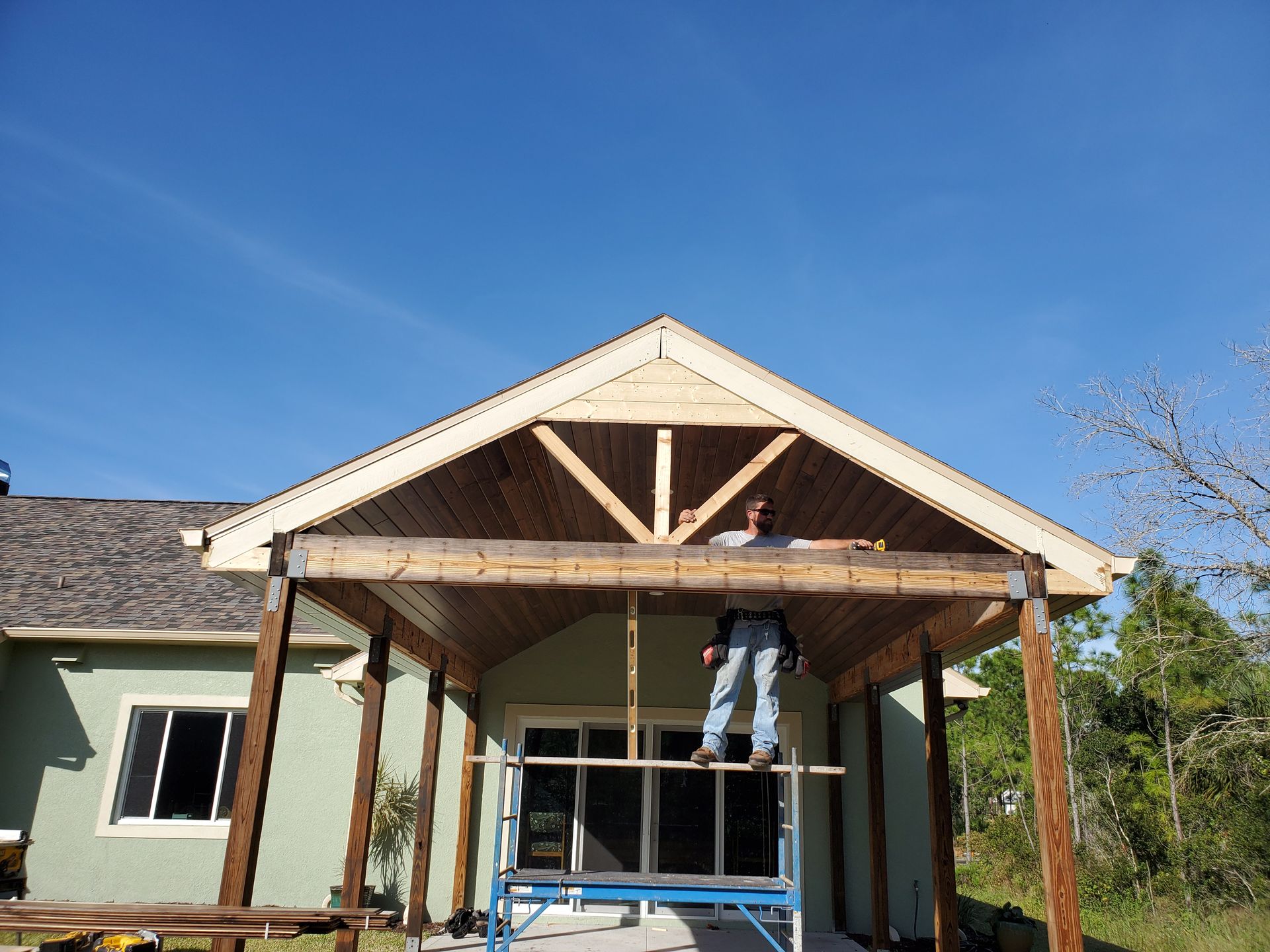 A construction worker stands on a blue metal scaffold, installing wooden trim on a large covered outdoor patio structure.