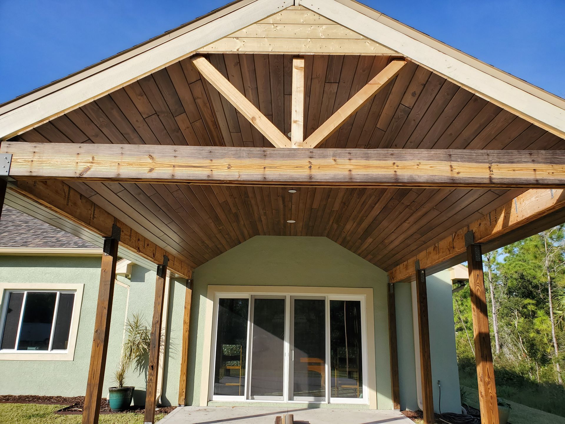 A light green house with a rustic wooden patio cover, featuring large timber beams and a vaulted plank ceiling.