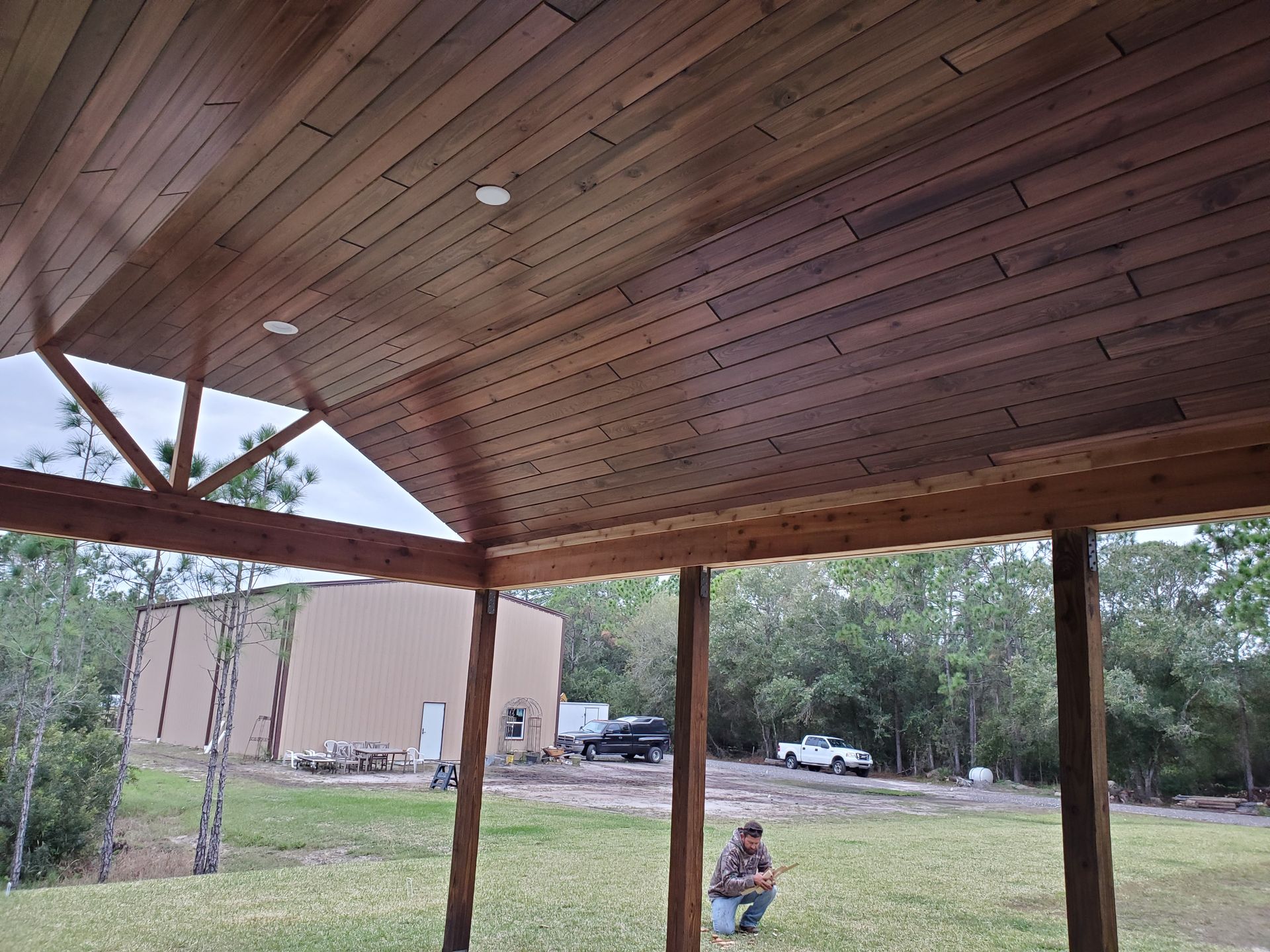 A person crouches on a lawn in front of a covered porch with a high, vaulted wooden ceiling.