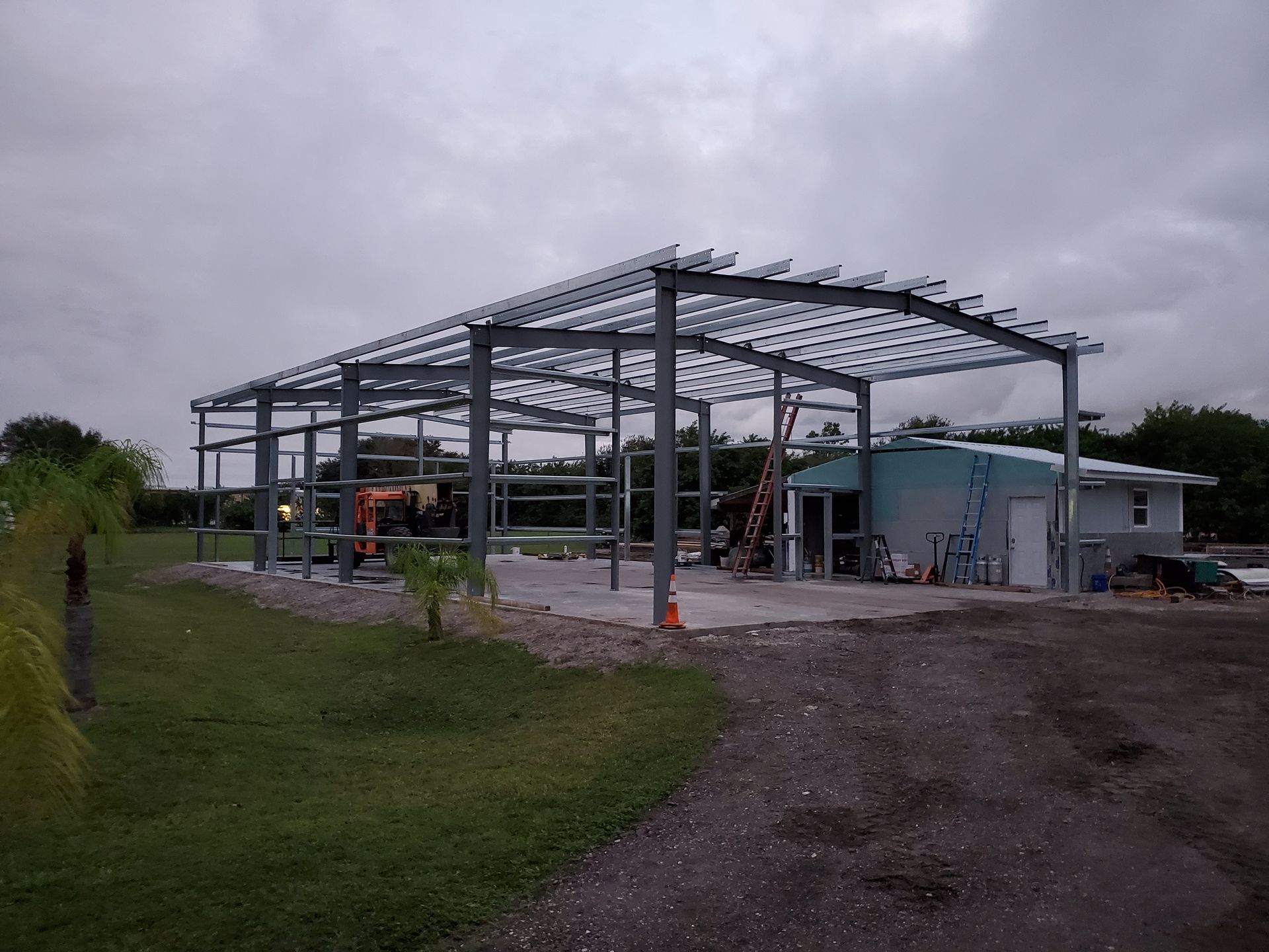 A steel building frame under construction outdoors, with a gravel lot and a small existing house nearby.