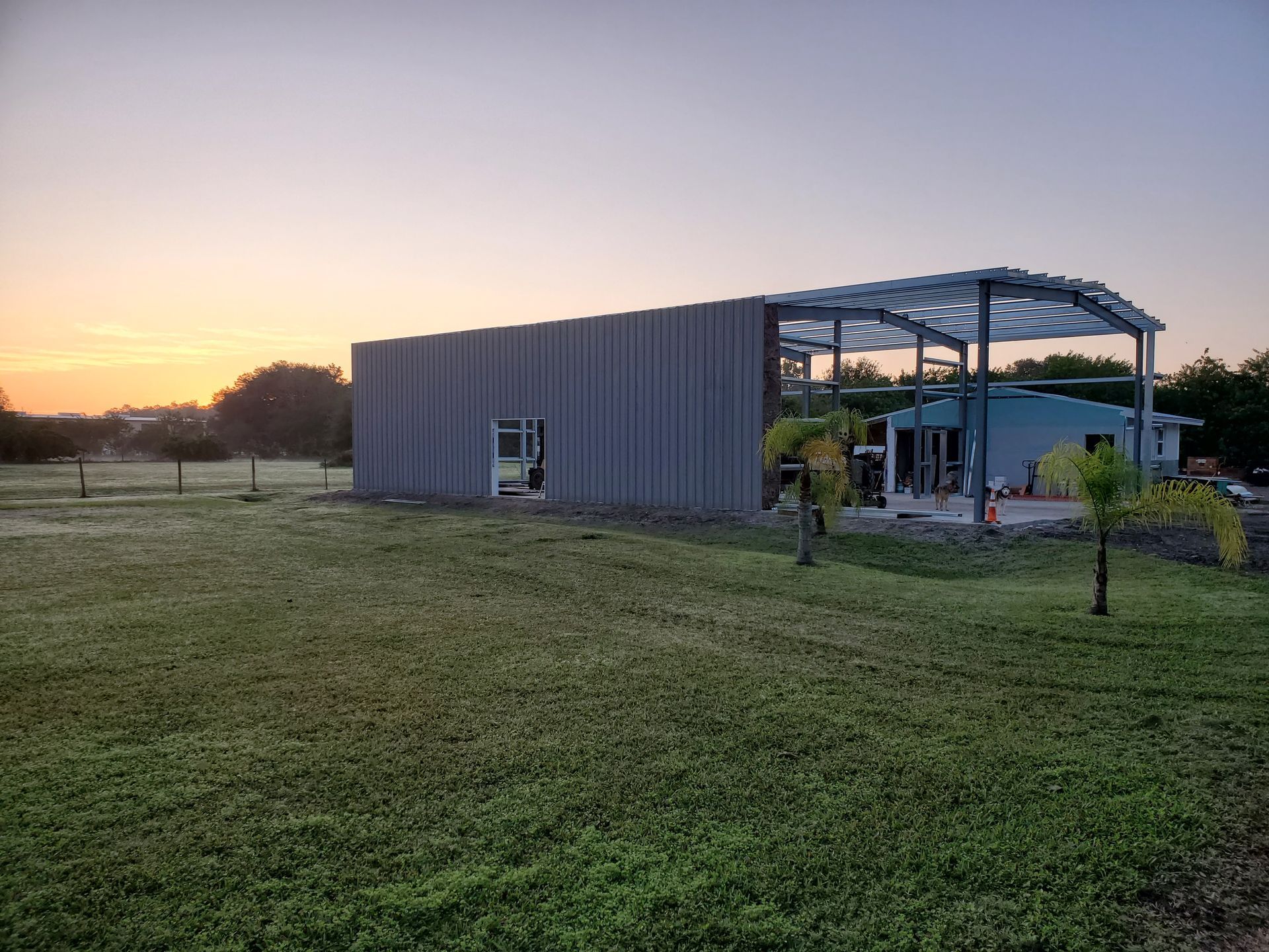 A large metal building under construction sits in a grassy field during a golden sunrise.