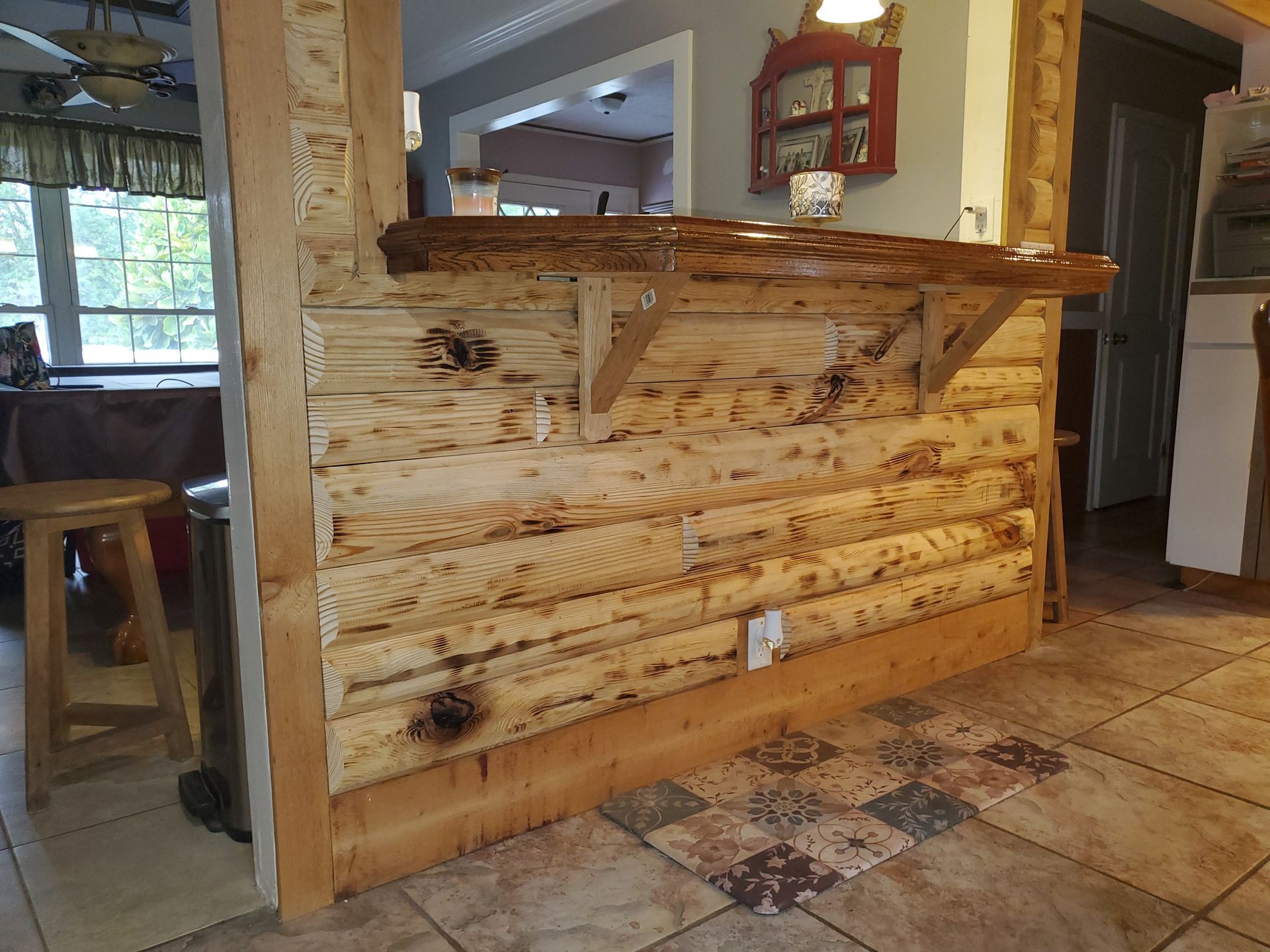 A rustic log-style breakfast bar with a wooden countertop and support brackets in a kitchen with tiled flooring.
