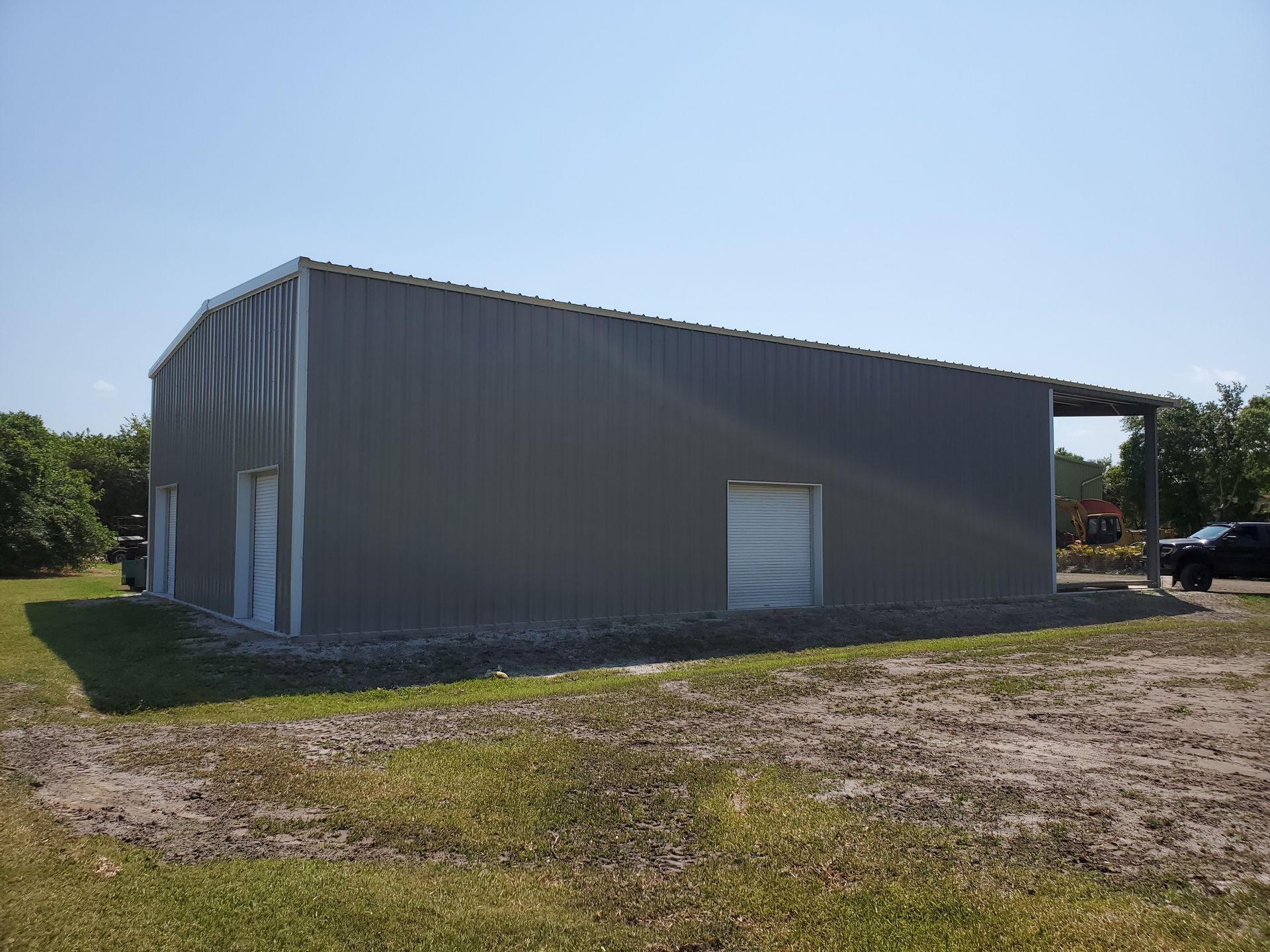 A large, gray metal warehouse building with a side canopy, situated on a grassy plot under a clear blue sky.