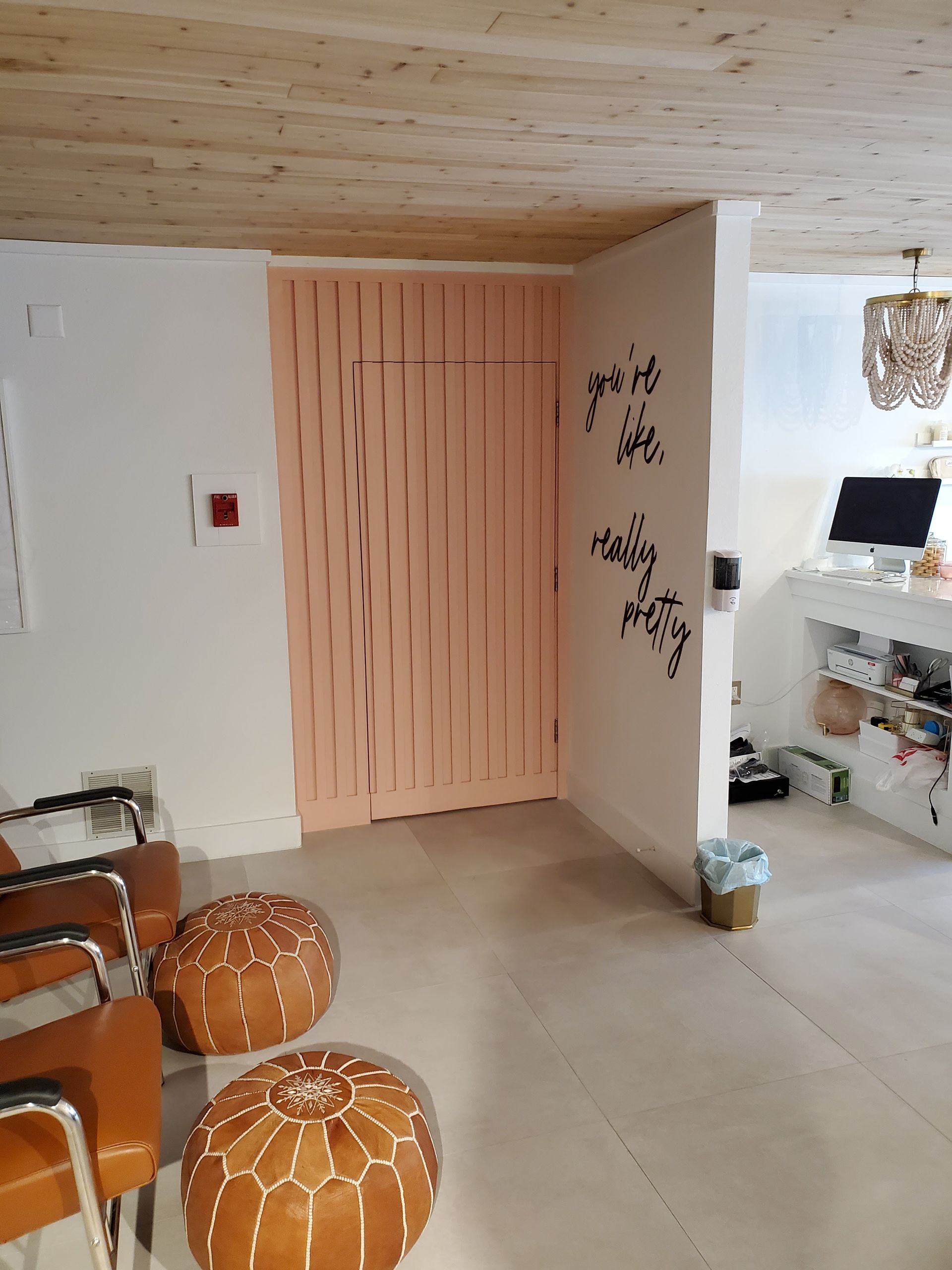 A modern waiting area with tan leather chairs, two round patterned poufs, a light-pink slatted door, and a white interior.