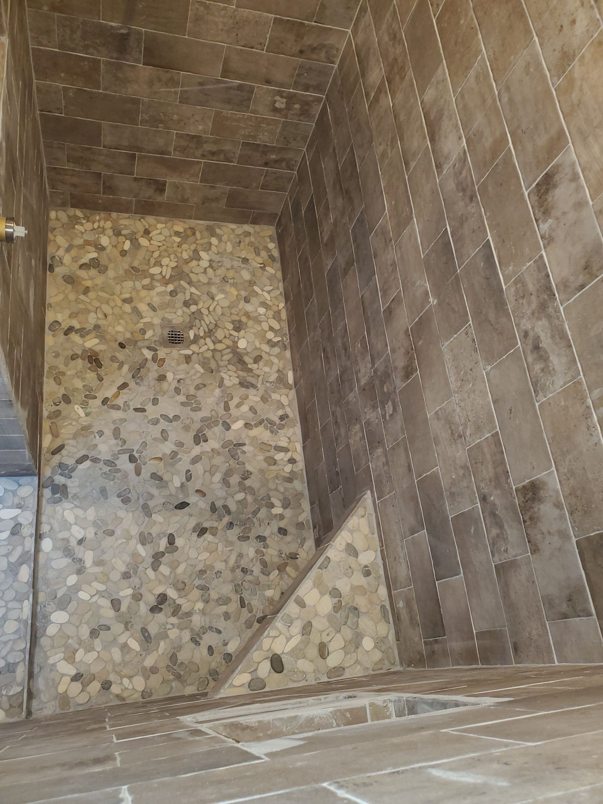 A shower interior under construction with brown rectangular wall tiles and a pebble-stone floor.