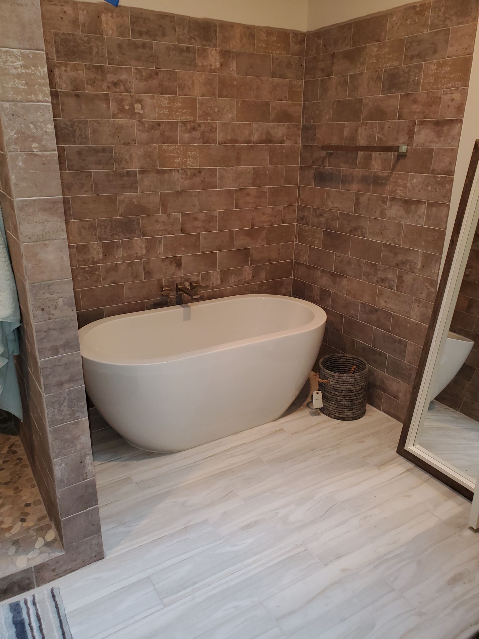 A modern white freestanding bathtub placed against a rustic brick wall in a bathroom with light wood-look tiled floors.