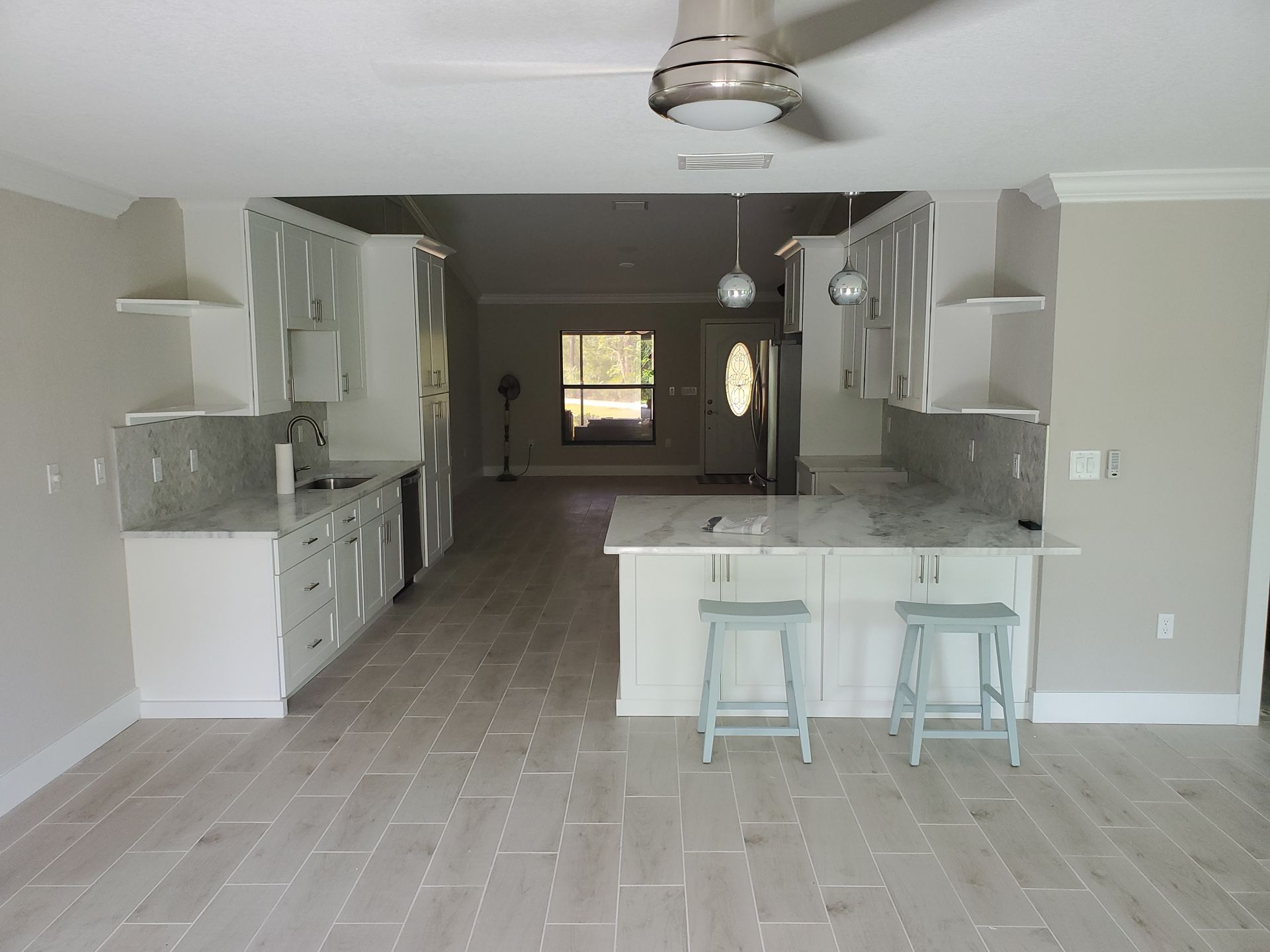A bright, modern kitchen featuring white cabinets, marble countertops, an island with two stools, and light tile flooring.