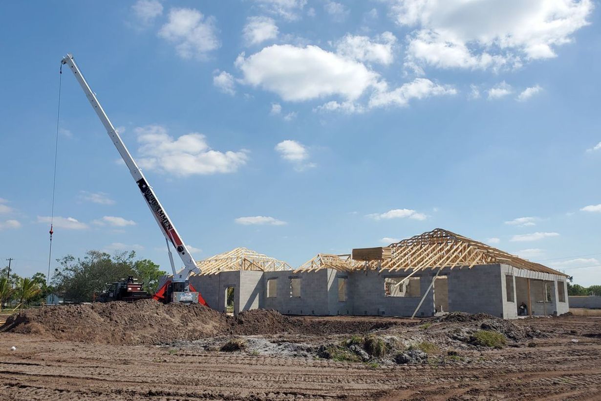 A crane stands beside a house under construction with exposed wooden roof trusses against a blue, partly cloudy sky.