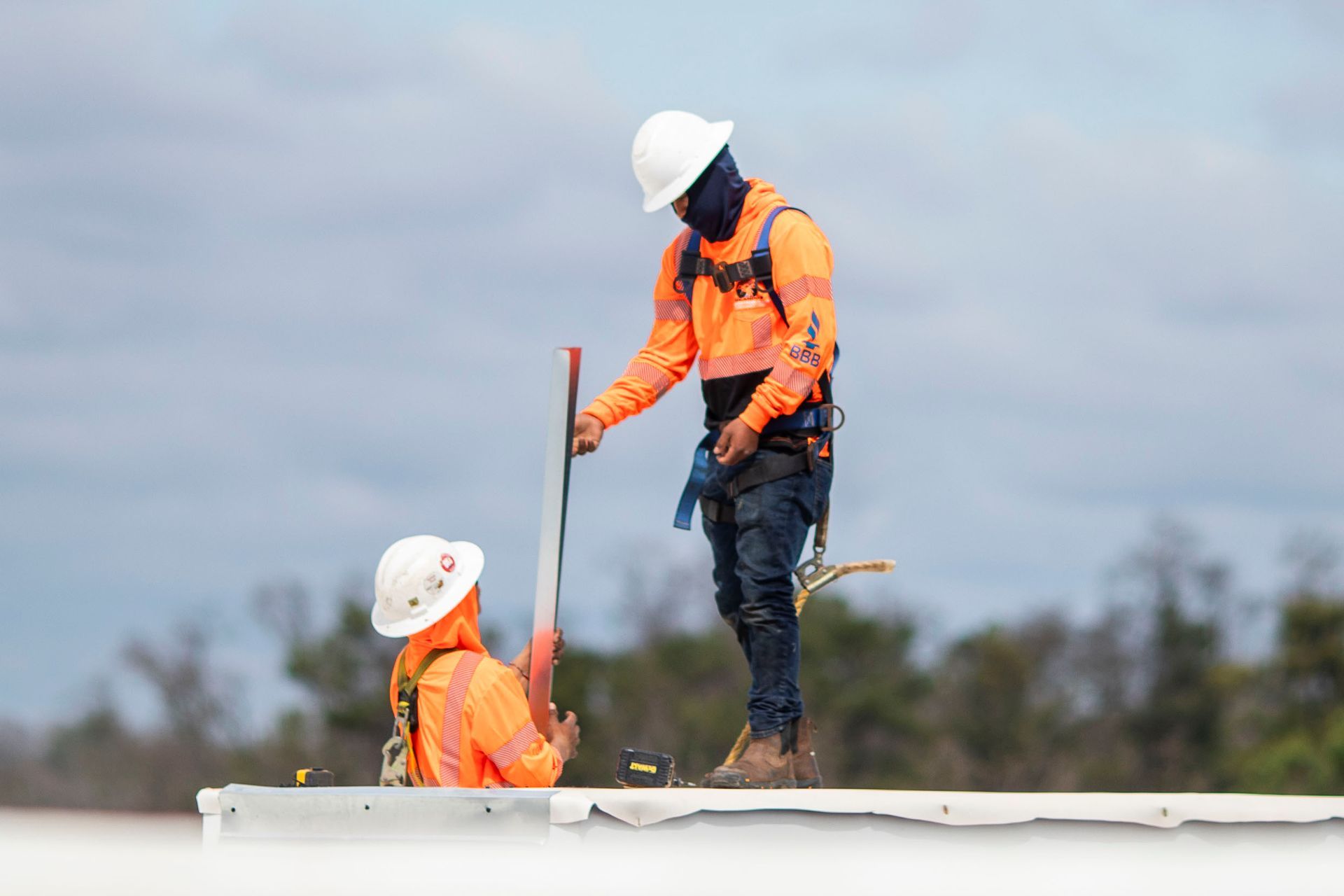 Two construction workers in hard hats and orange vests measure on a rooftop.