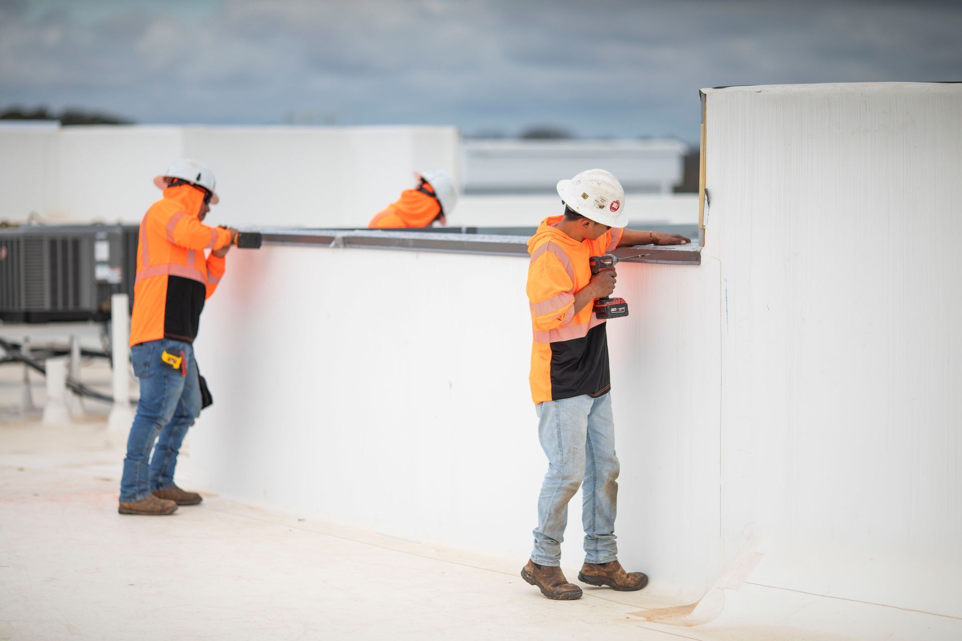 Workers in orange safety vests and white hard hats inspecting a white wall on a rooftop.