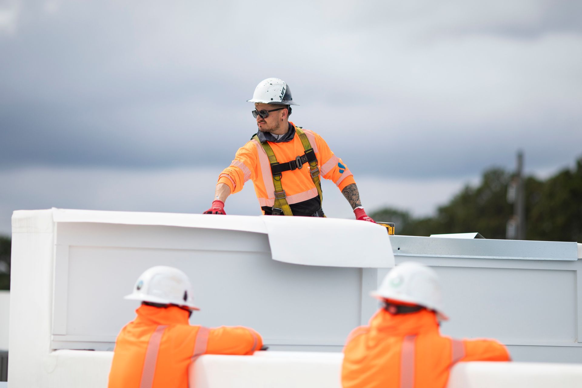 Workers in orange safety gear and white helmets moving large white panels at a construction site