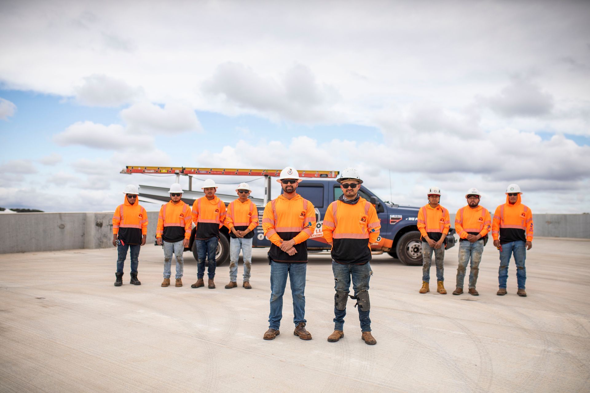 Workers in orange safety gear stand on a rooftop under a cloudy sky.