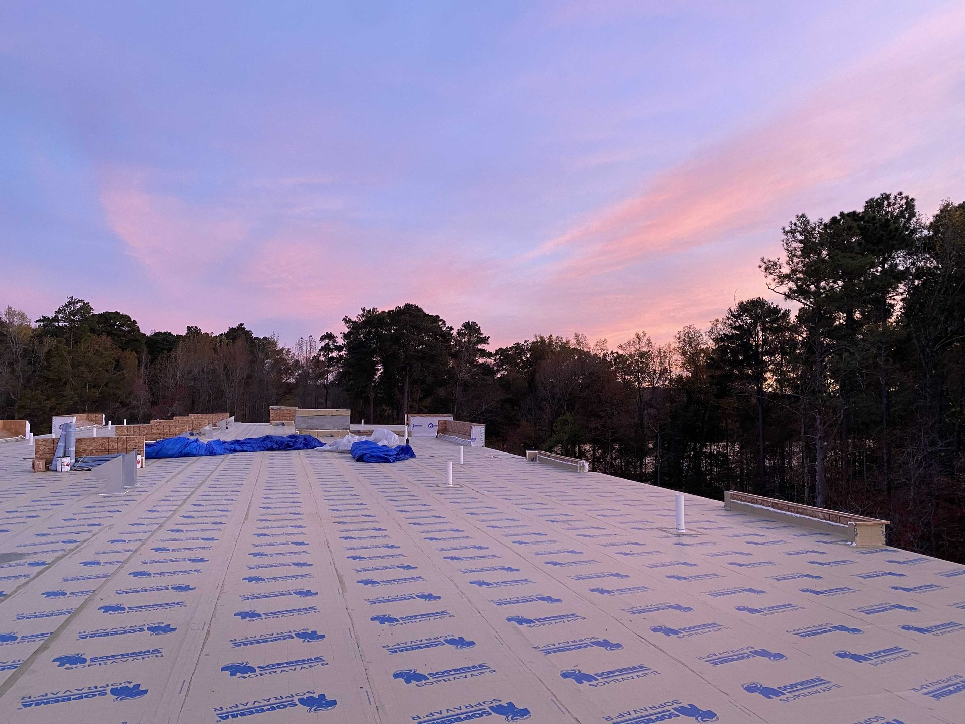 Flat rooftop with blue protective sheeting at sunset under a pink sky, trees in the background