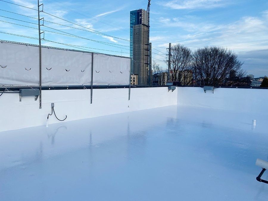 Outdoor frozen ice rink beside a tall building under a clear blue sky, with snow-covered edges and netting.