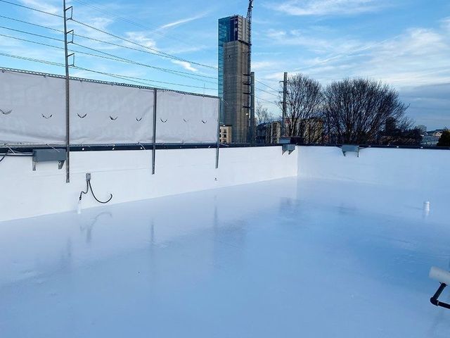 Empty outdoor ice rink with snow-covered surface, fence, and a tall building under a blue sky