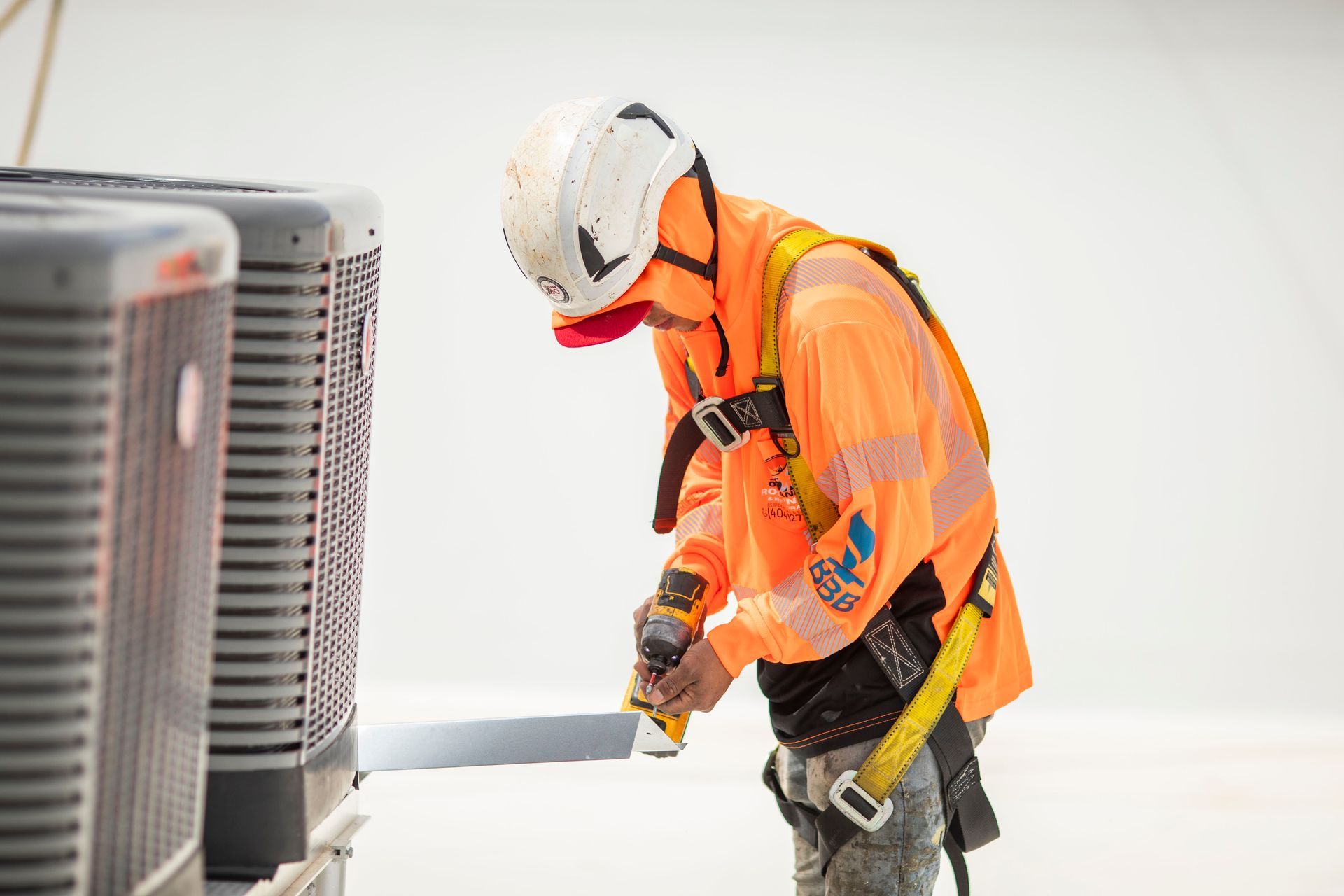 Worker in orange safety gear using a power drill to service outdoor HVAC units