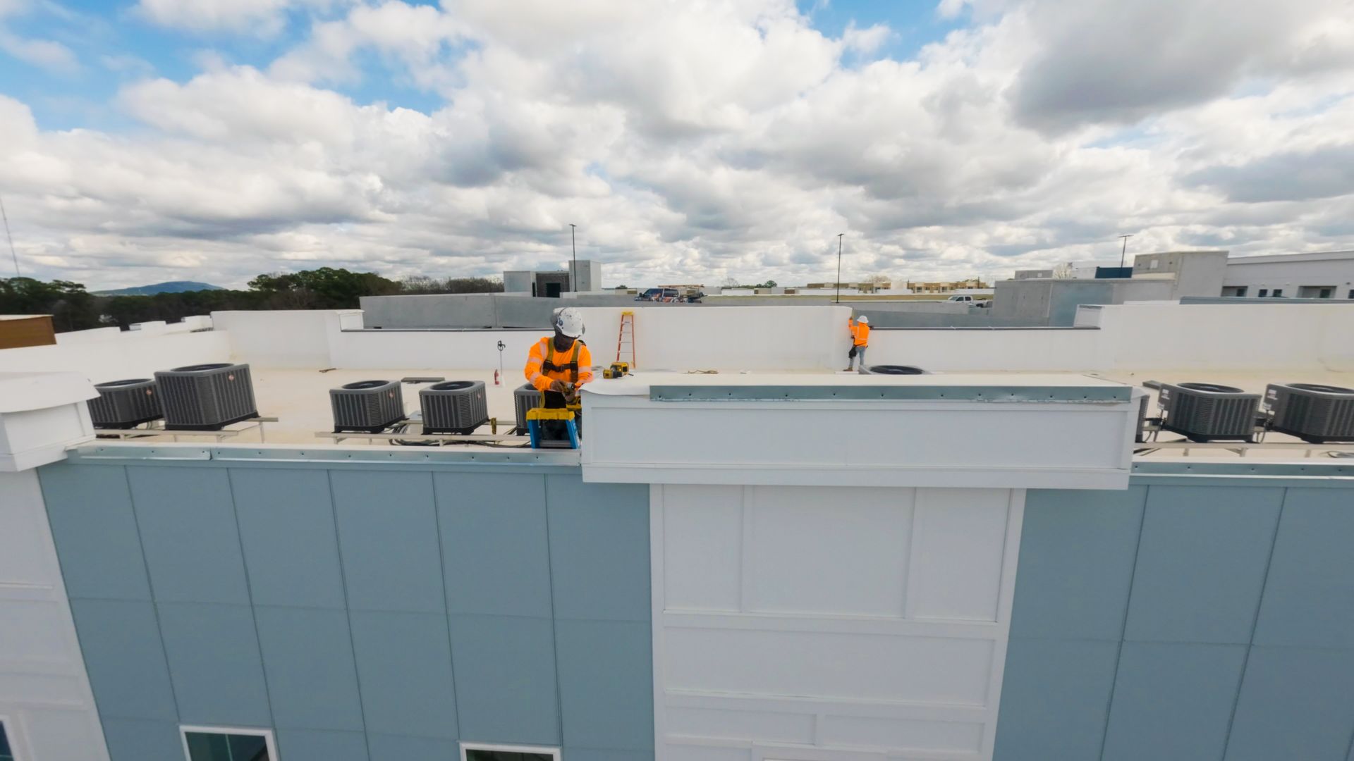 Rooftop workers in orange safety gear on a white building under a cloudy sky