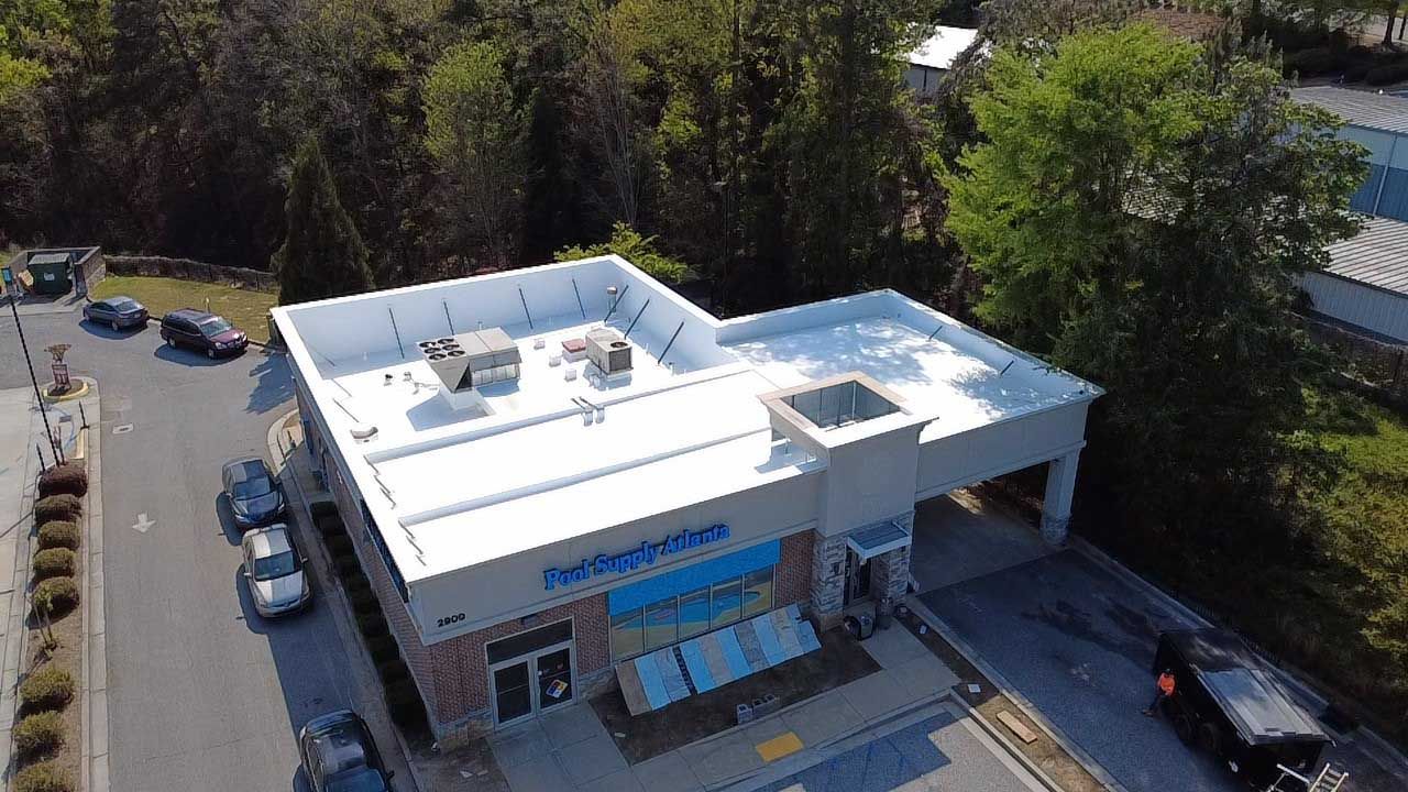 Aerial view of a white commercial building with blue signage and cars parked outside, surrounded by trees.