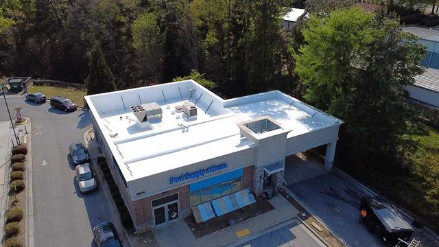 Aerial view of a white commercial building with a blue awning, parking lot, and surrounding trees.