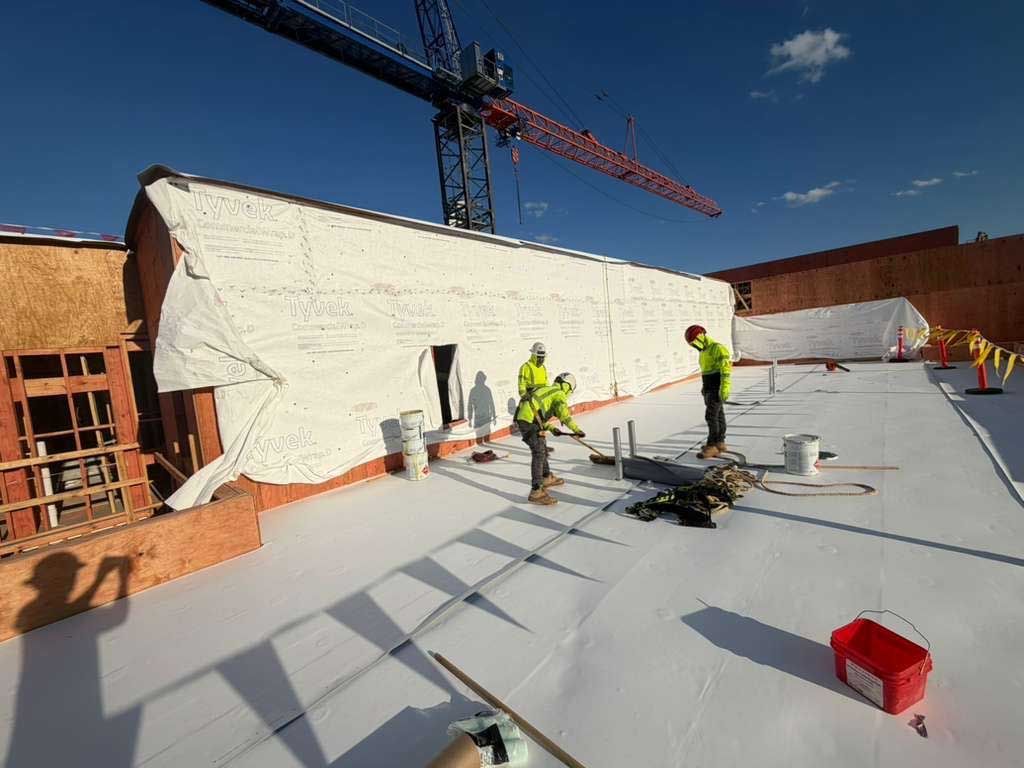Workers on a rooftop near a crane, with white roofing material and construction tools scattered around.