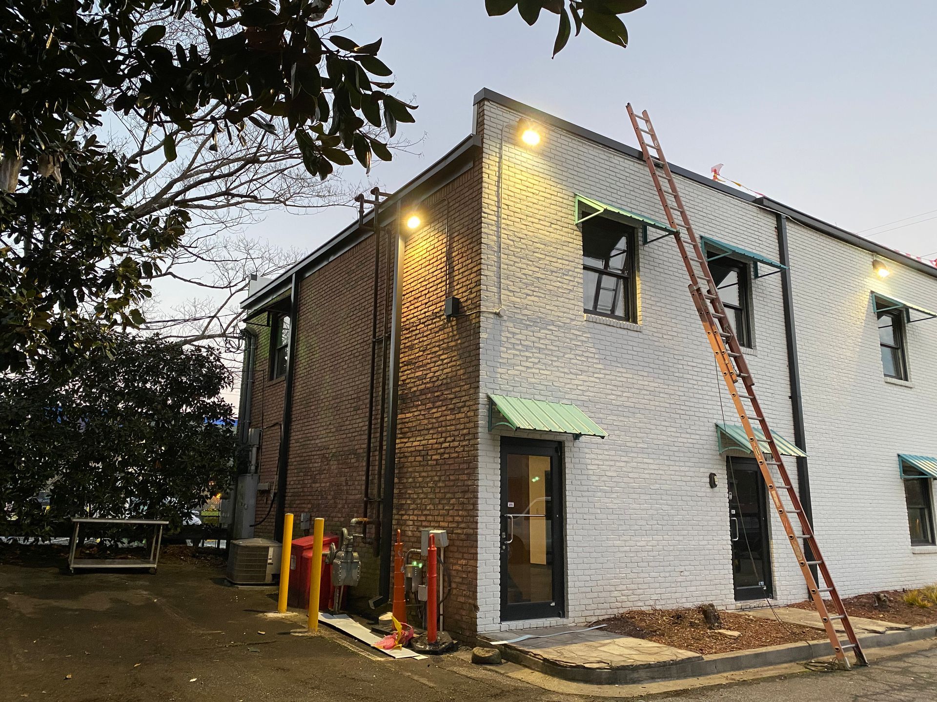 White brick building exterior with ladders, red pipes, and an open doorway at dusk