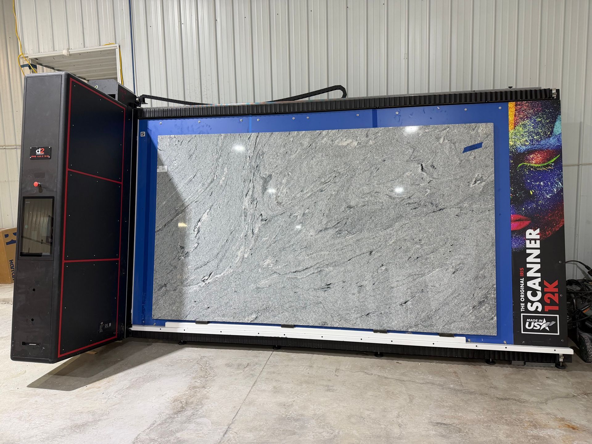 A large industrial stone slab scanner with a speckled gray granite slab mounted on its blue frame in a warehouse.