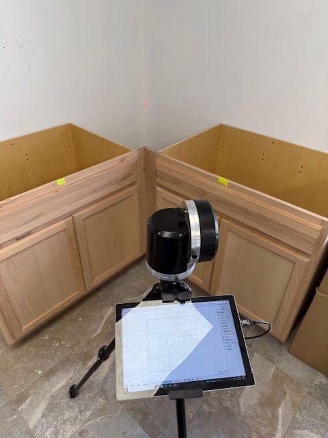 A 3D scanner on a tripod measuring cabinetry in a corner. A tablet displays a diagram.