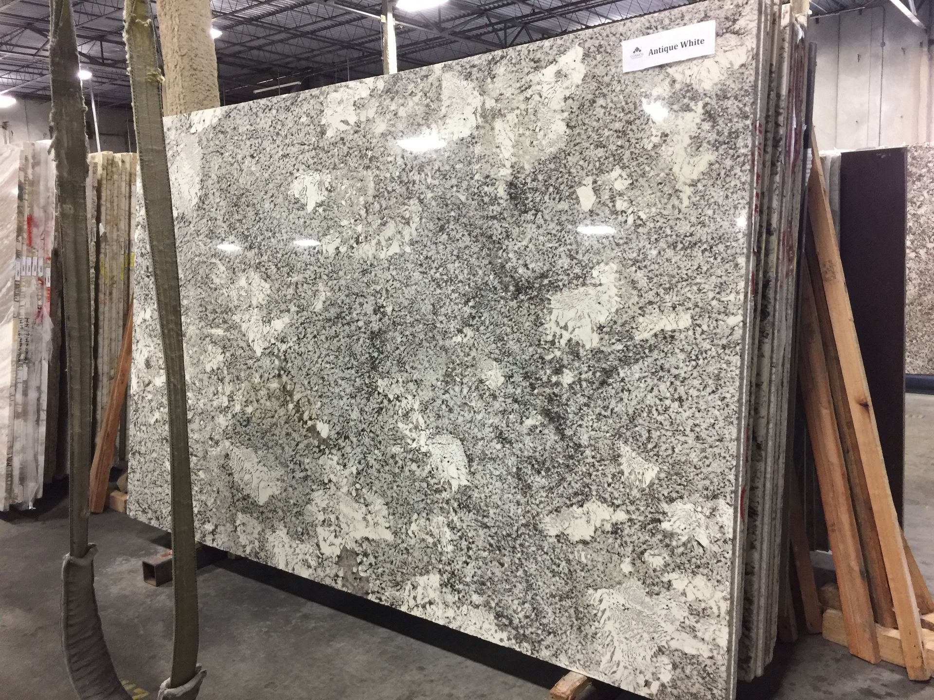Large granite slab with white, gray, and tan speckles, standing upright in a warehouse.