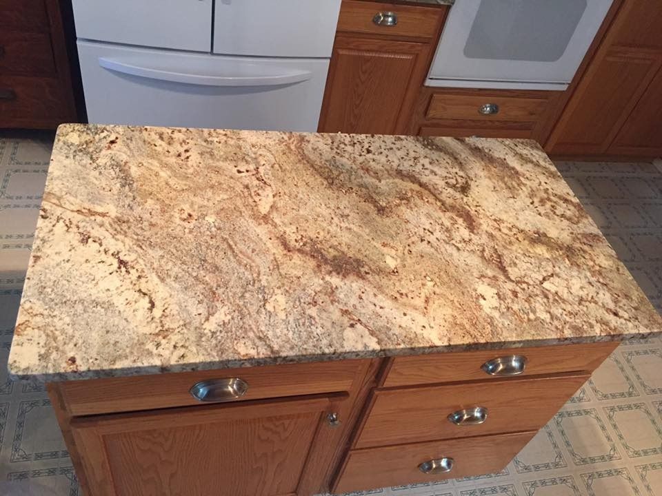 Granite countertop on a wooden kitchen island with cabinets and drawers, near a refrigerator and stove.