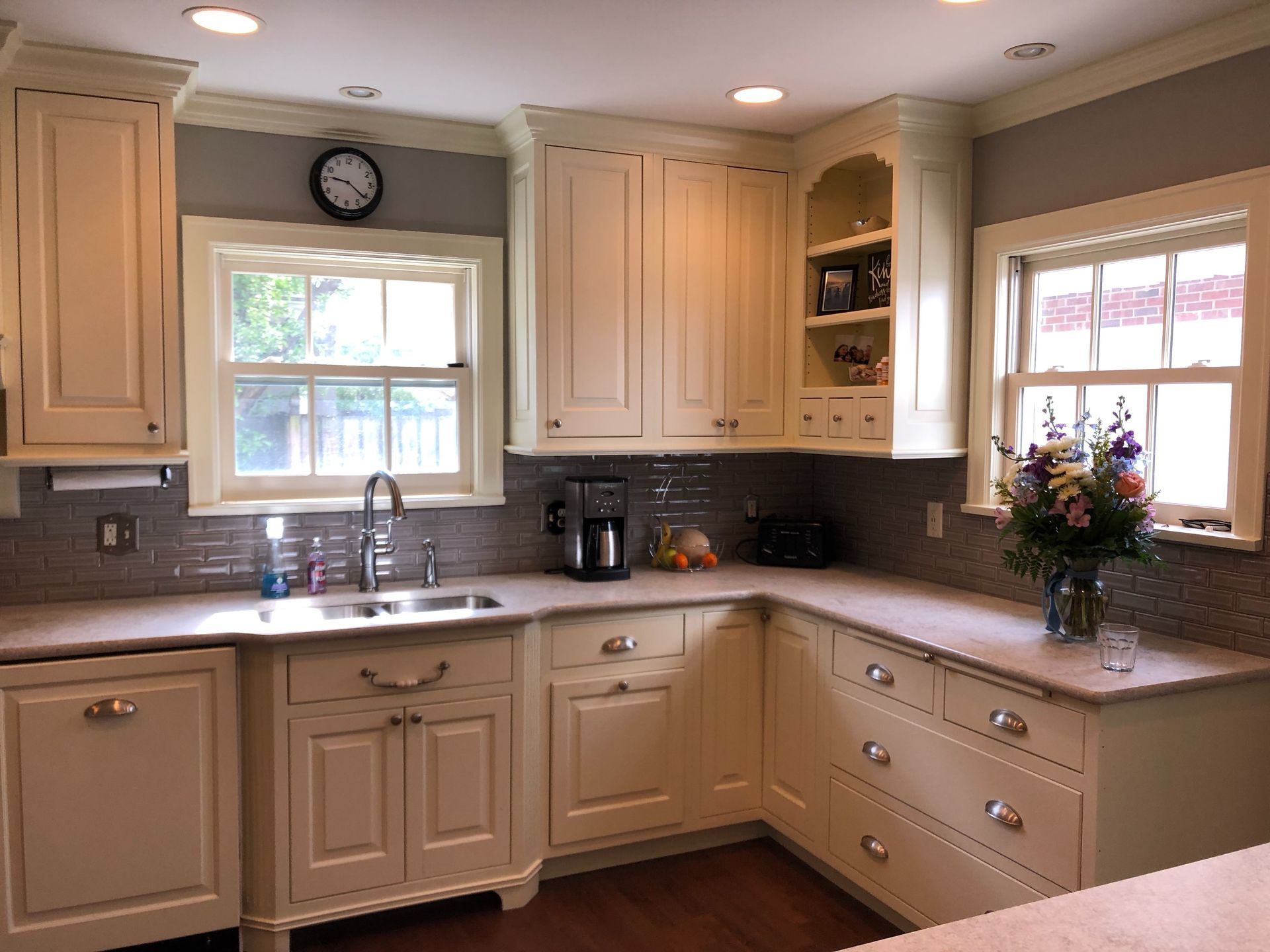 Cream-colored kitchen cabinets with gray countertops. Stainless steel sink, coffee maker, and colorful flowers.