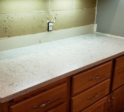 Kitchen countertop with brown cabinets and damaged wall. Electrical outlet visible.