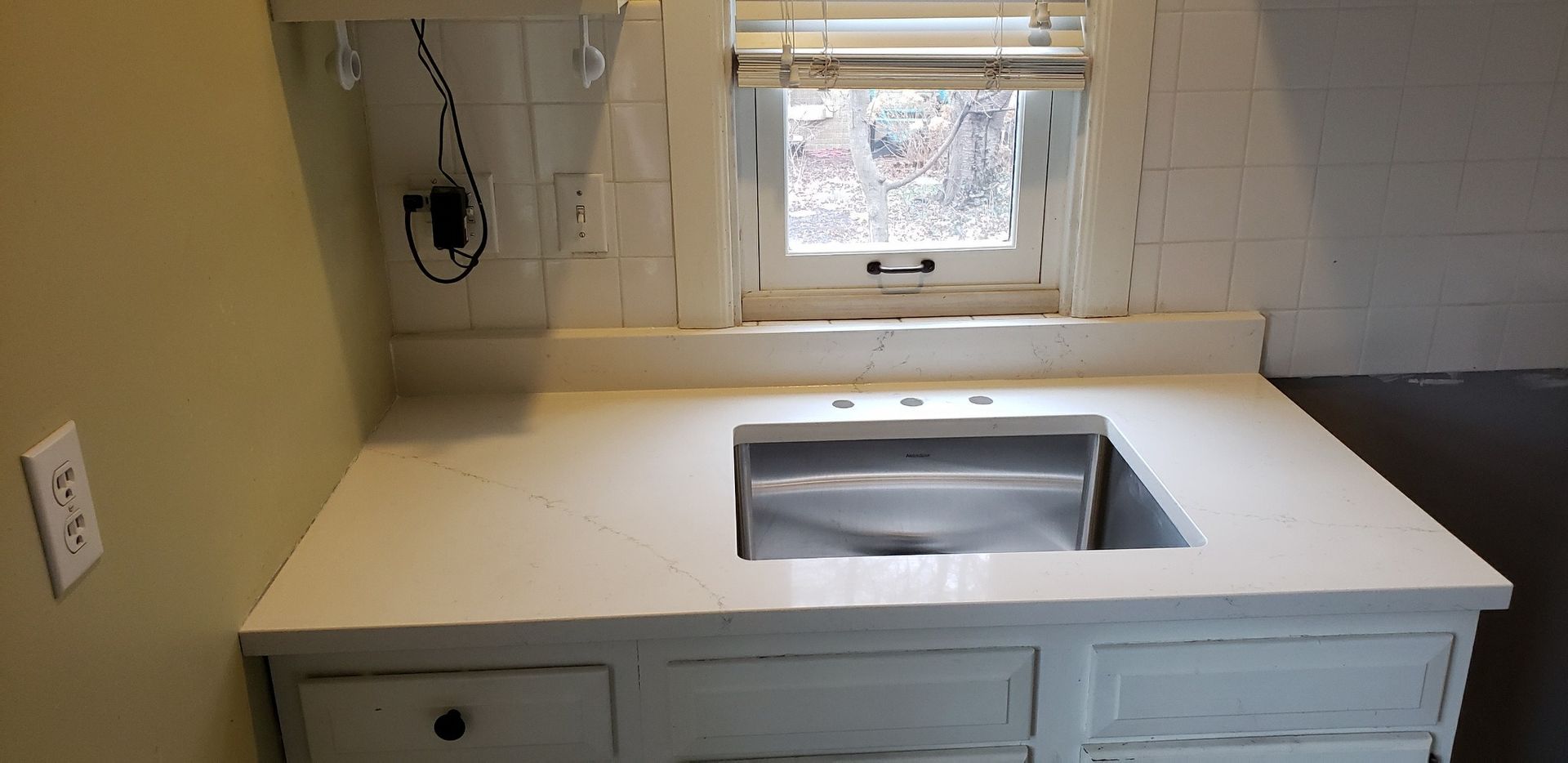 White kitchen sink with a window above, set against a backdrop of white tile and a pale yellow wall.