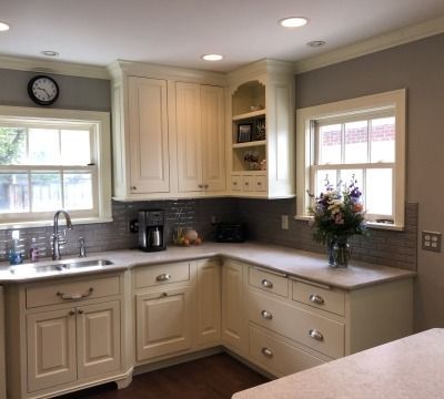 Cream-colored kitchen cabinets and counters in a corner layout. Windows, sink, and appliances are visible against gray walls.