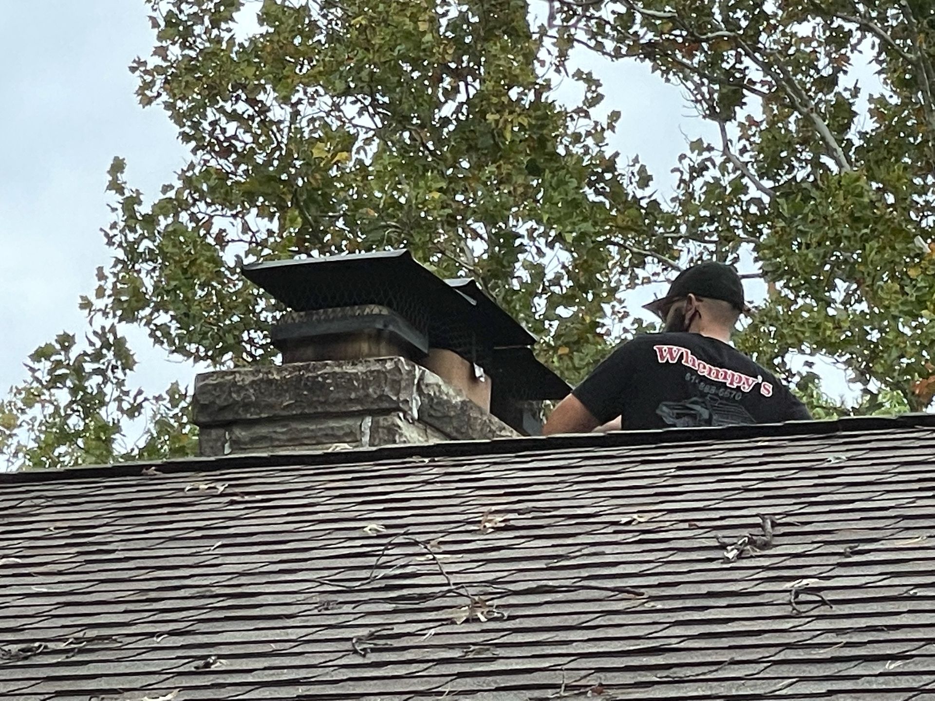 Person installing a black chimney cap on a brick chimney atop a shingled roof.