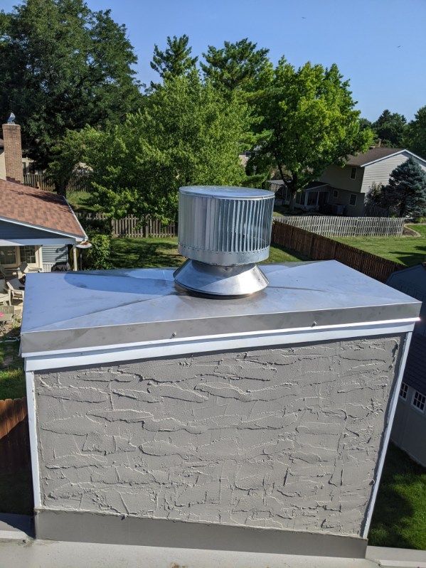 Gray stucco chimney with a metal cap and rotating vent against a background of trees and sky.