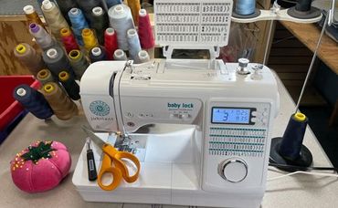 White sewing machine on a table with colorful thread spools, scissors, and a pink pin cushion nearby