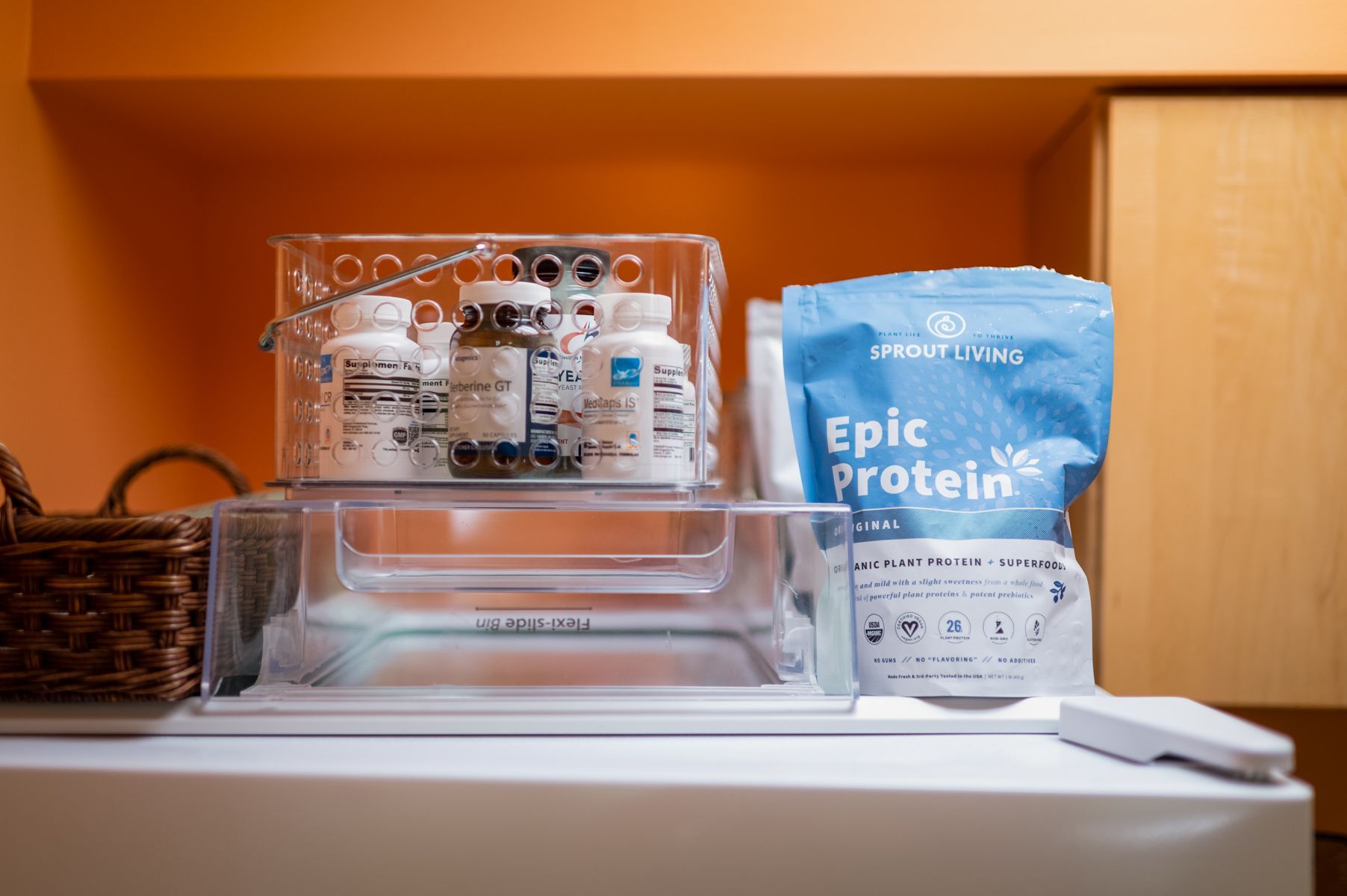 Supplements, protein powder, and small storage container on a counter. Orange background.