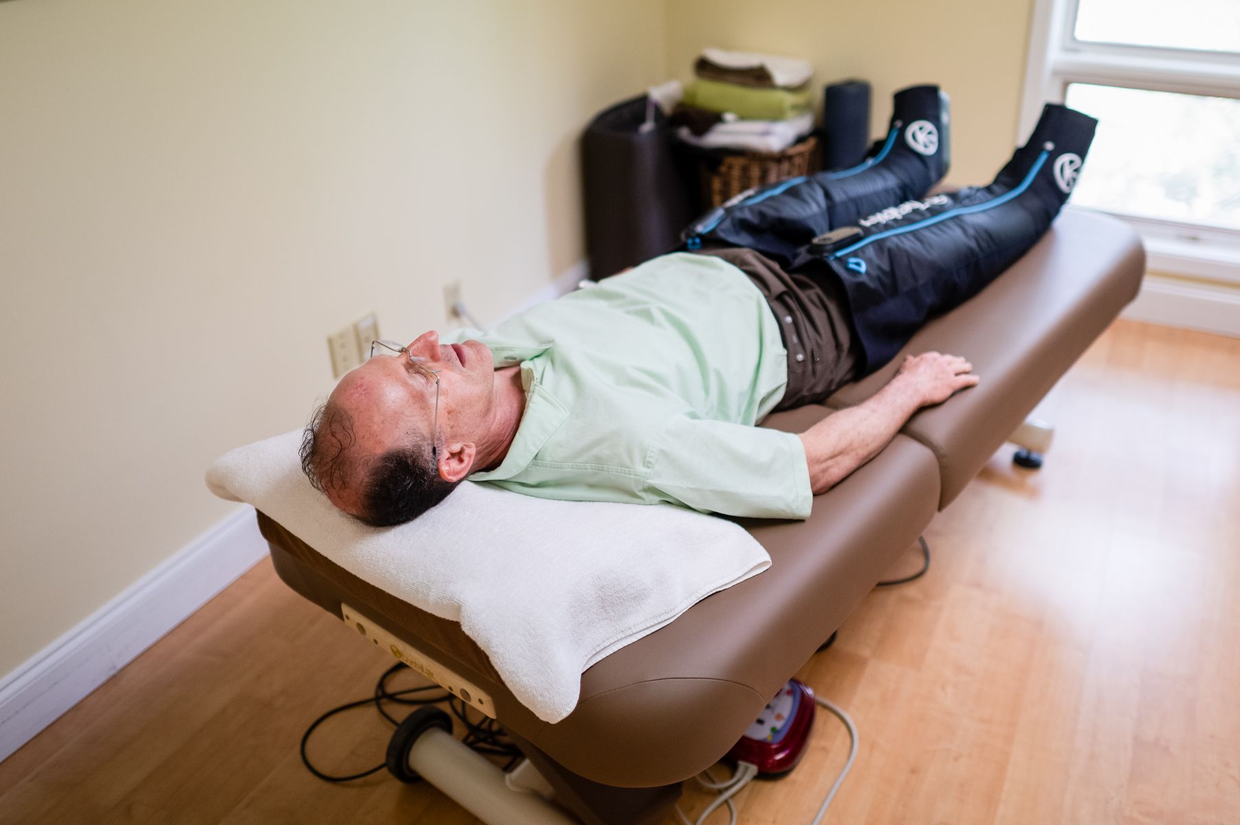 Man lying on a massage table with leg compression boots. Indoors, light-skinned, pale green shirt.