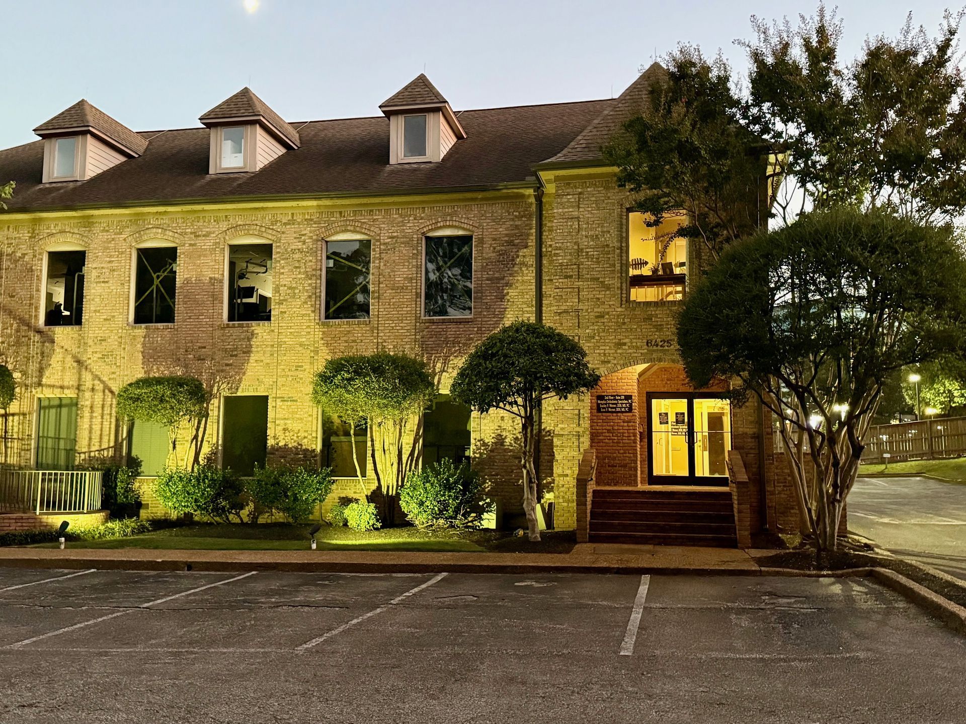 Two-story brick building with dormers. Landscaped with lights, trees, and a dark parking lot.
