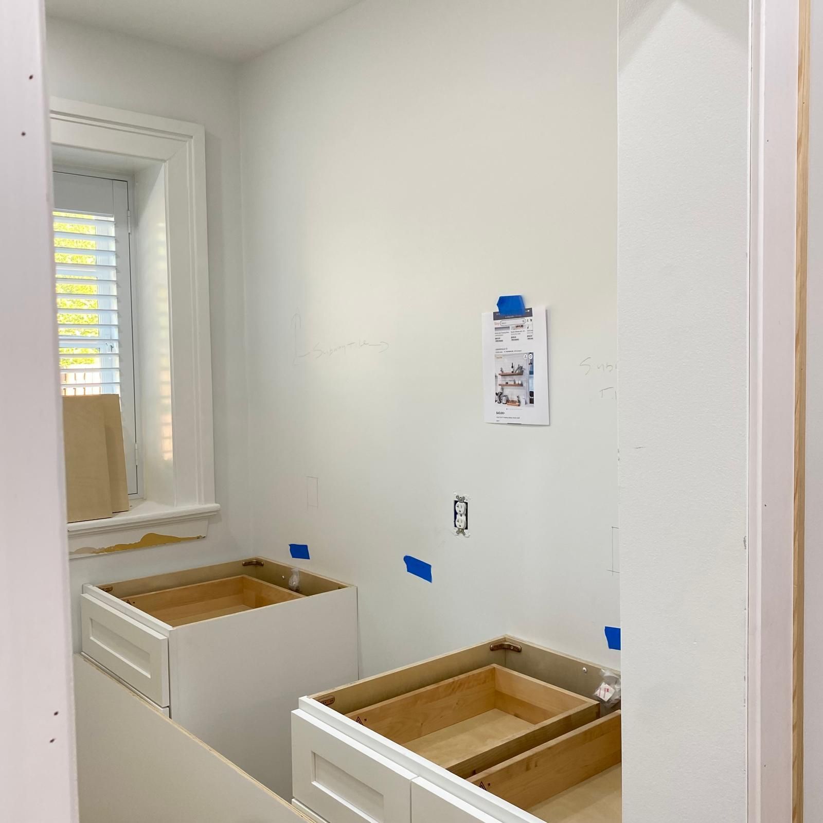 Small white bathroom under renovation with two unfinished vanity cabinets and blue painter’s tape on the walls