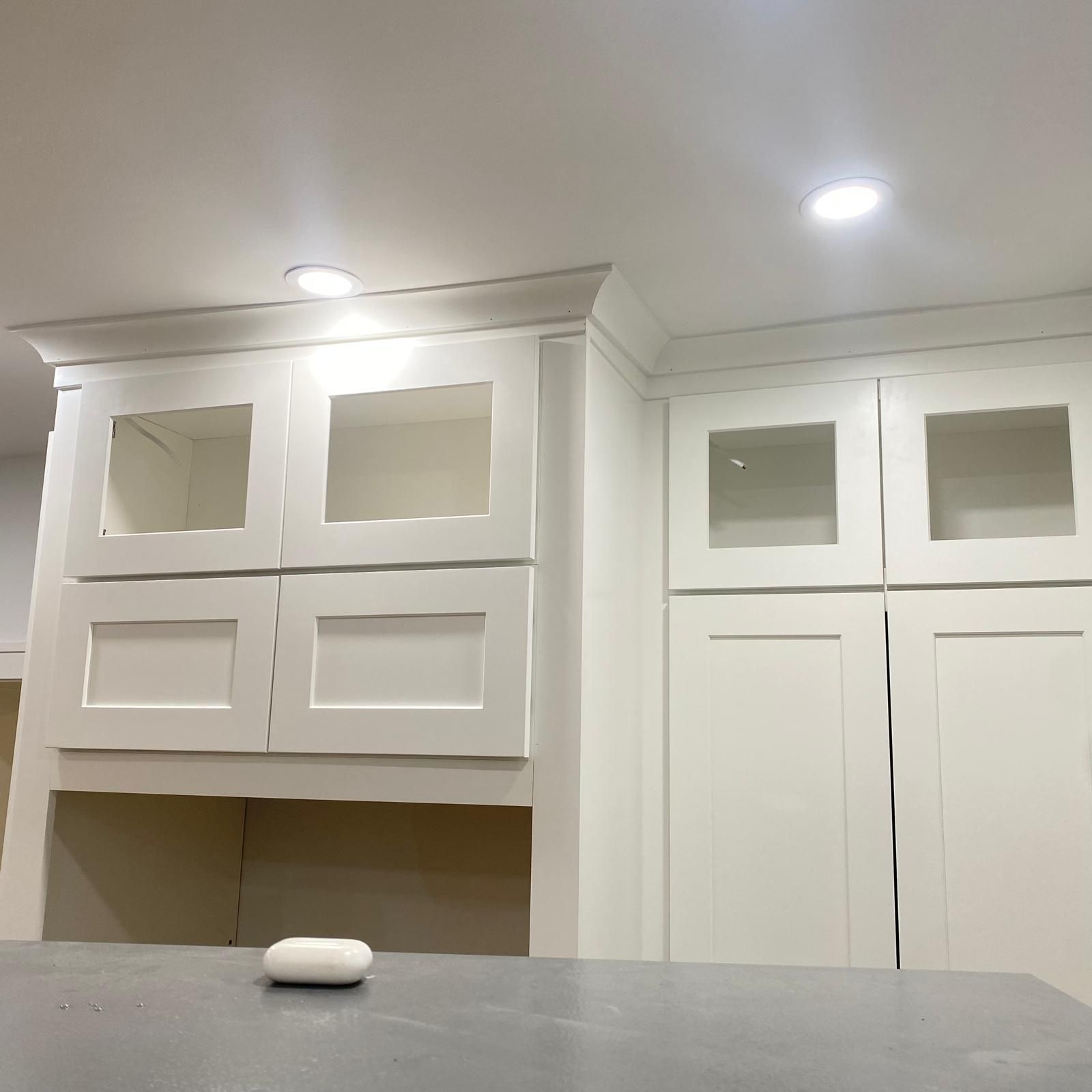 Bright white kitchen cabinets with glass doors above a countertop under recessed ceiling lights.