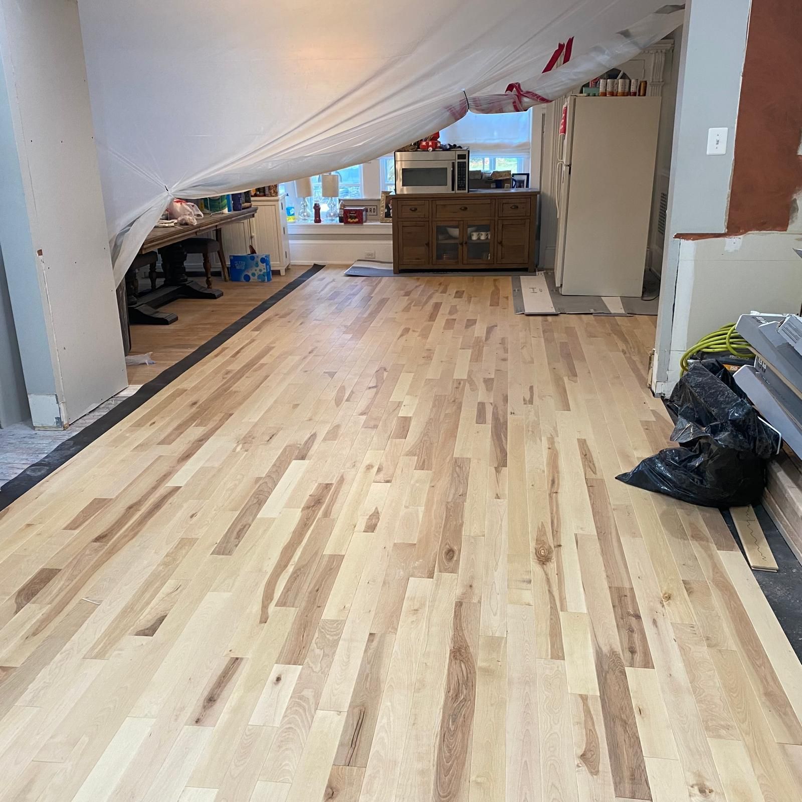 Unfinished room with new hardwood flooring, draped plastic barrier, and furniture at the back.