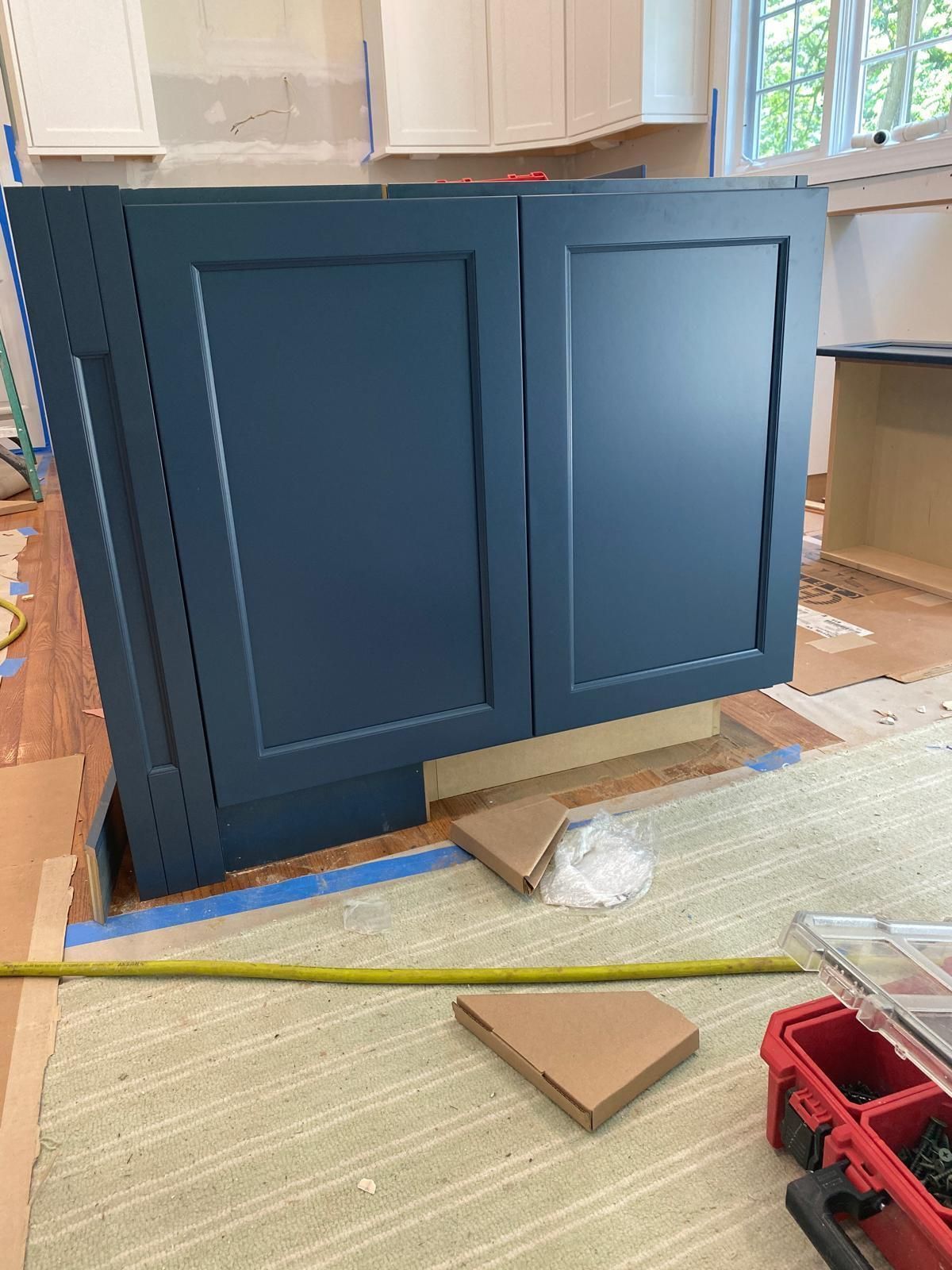 Blue kitchen cabinet being installed on a floor, with tools and packing materials nearby.