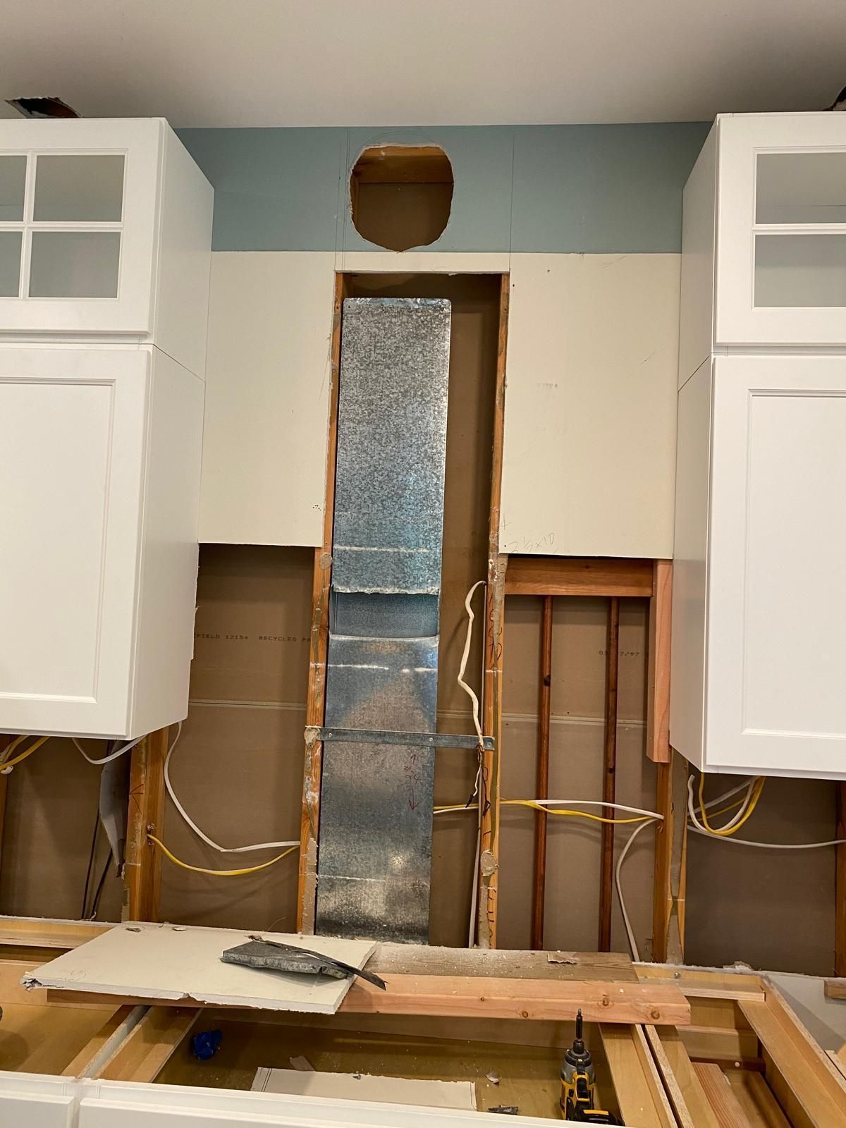 Kitchen under renovation with open wall studs, exposed wiring, and white cabinets on both sides