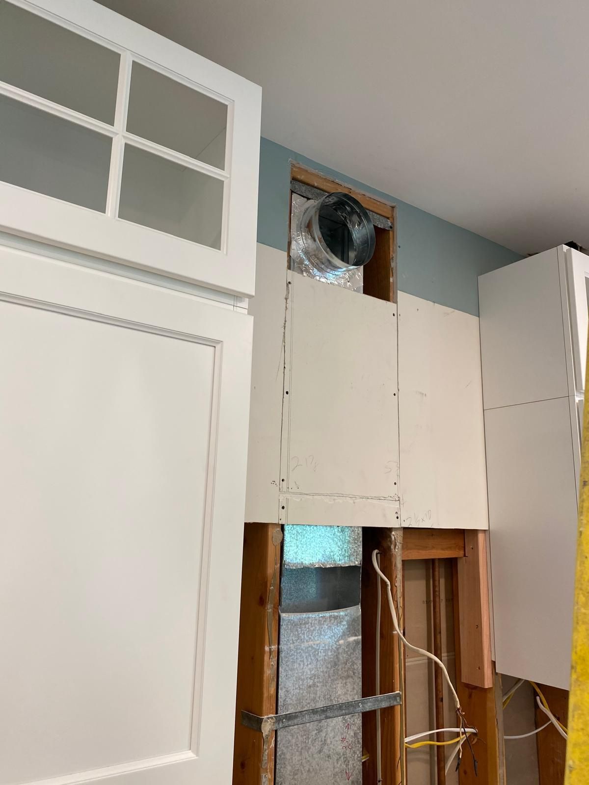 Partially open wall with exposed insulation, ductwork, and loose wires beside white cabinets in a room