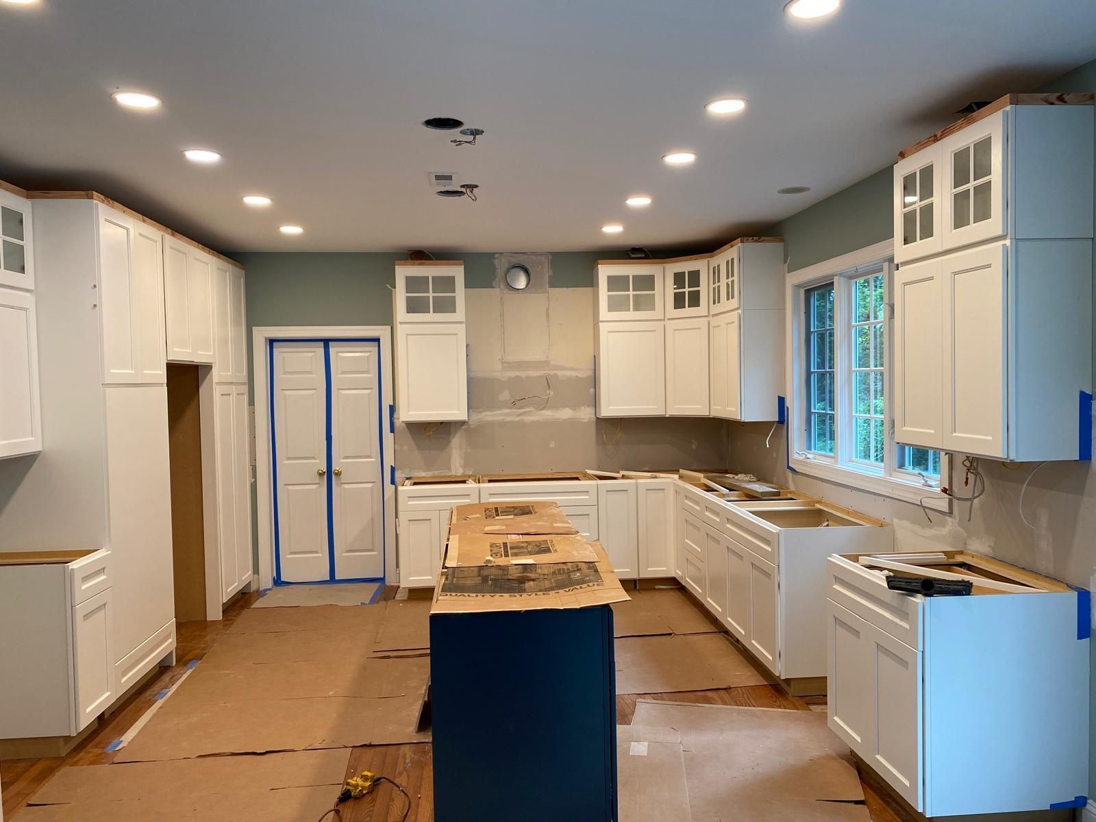 Unfinished kitchen with white cabinets, blue island, and cardboard-covered floor under recessed lights