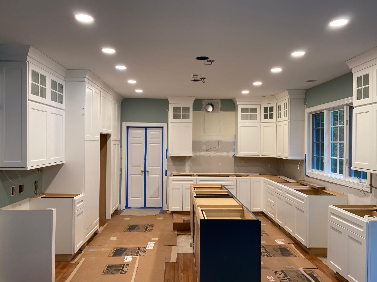 Bright white kitchen under renovation, with cabinets installed and unfinished floor and counters.