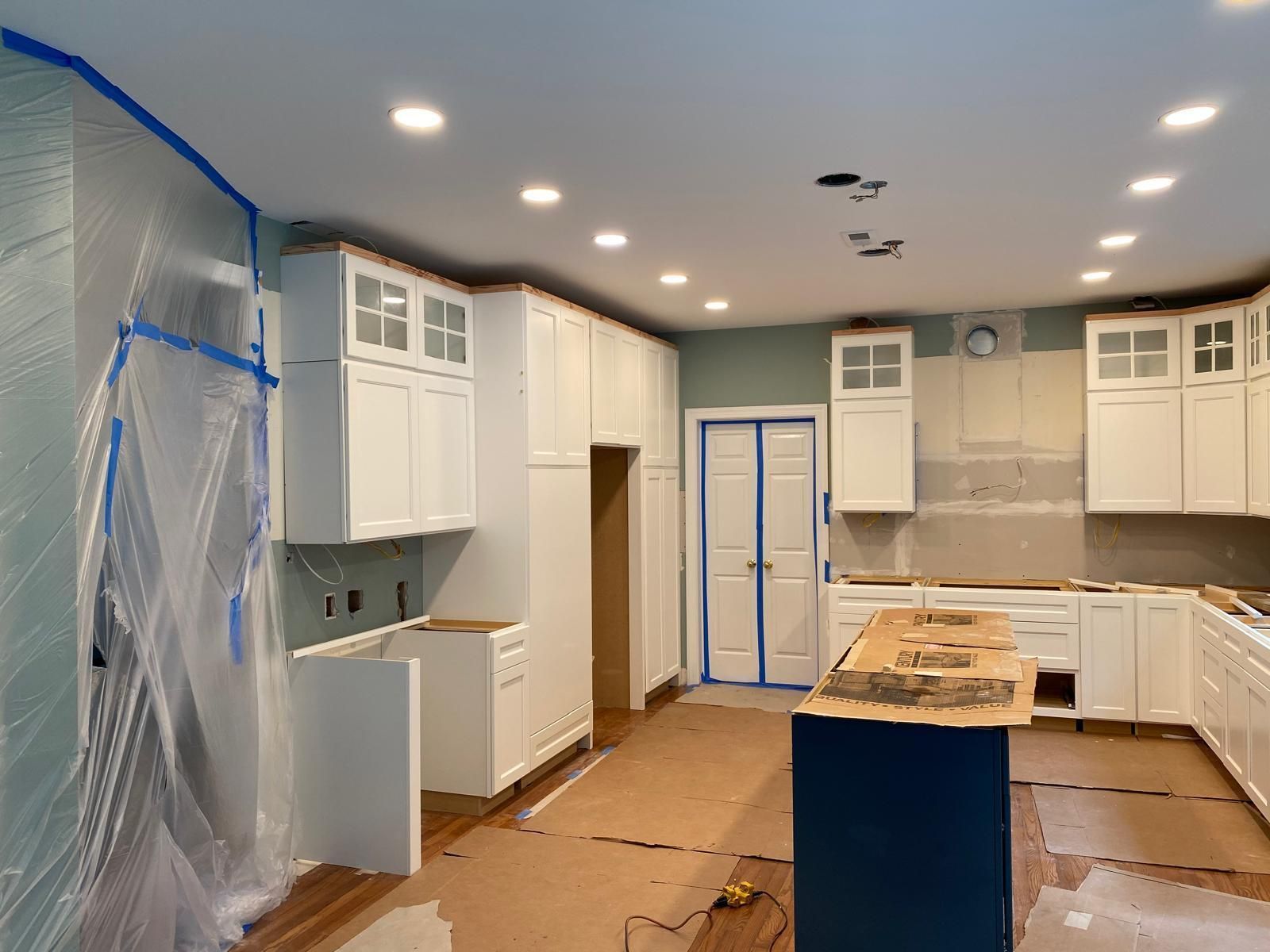 Kitchen under renovation with white cabinets, blue tape, plastic-covered wall, and an unfinished island countertop