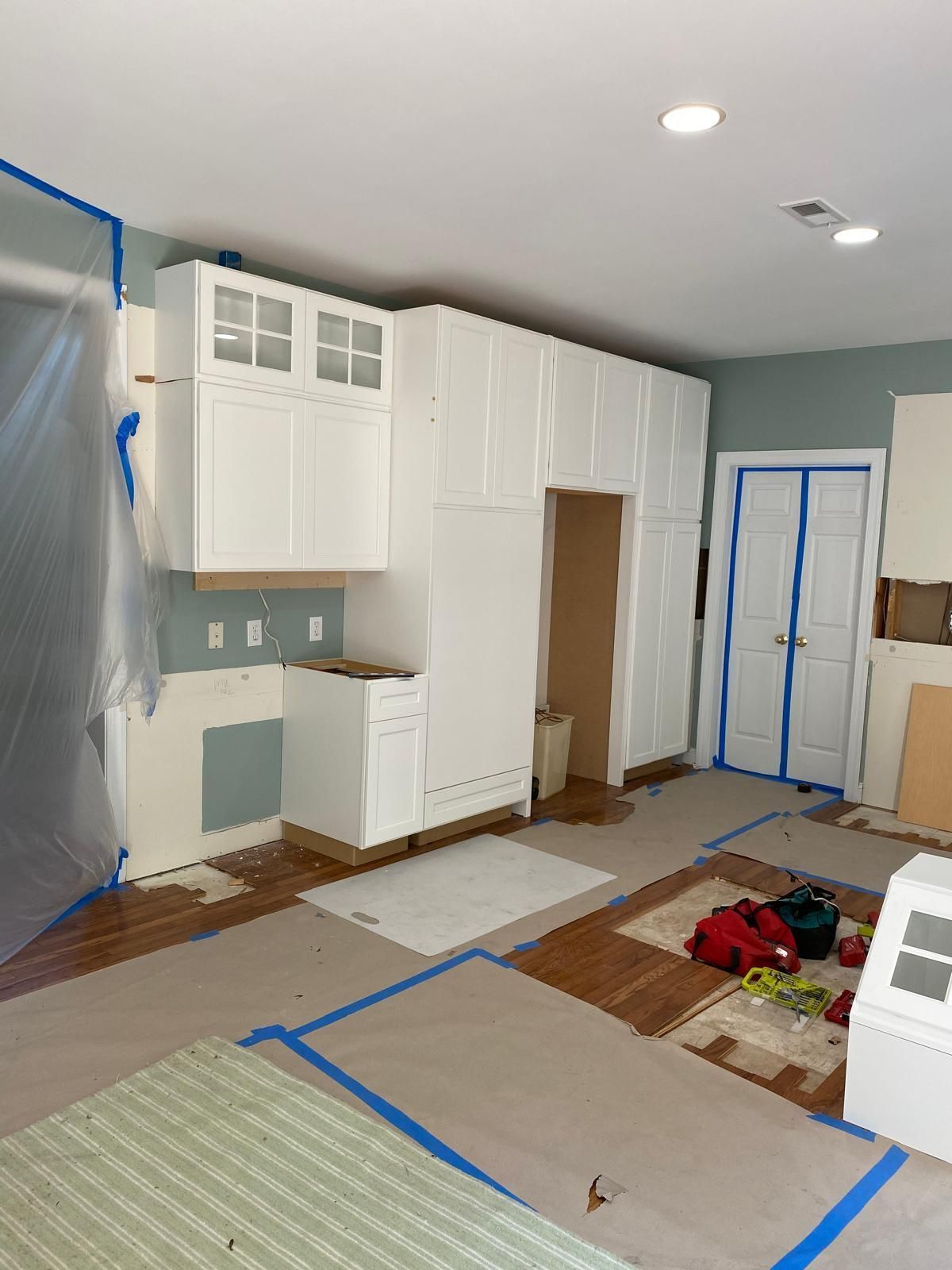 Unfinished kitchen with white cabinets, blue painter’s tape, and covered floors during renovation.