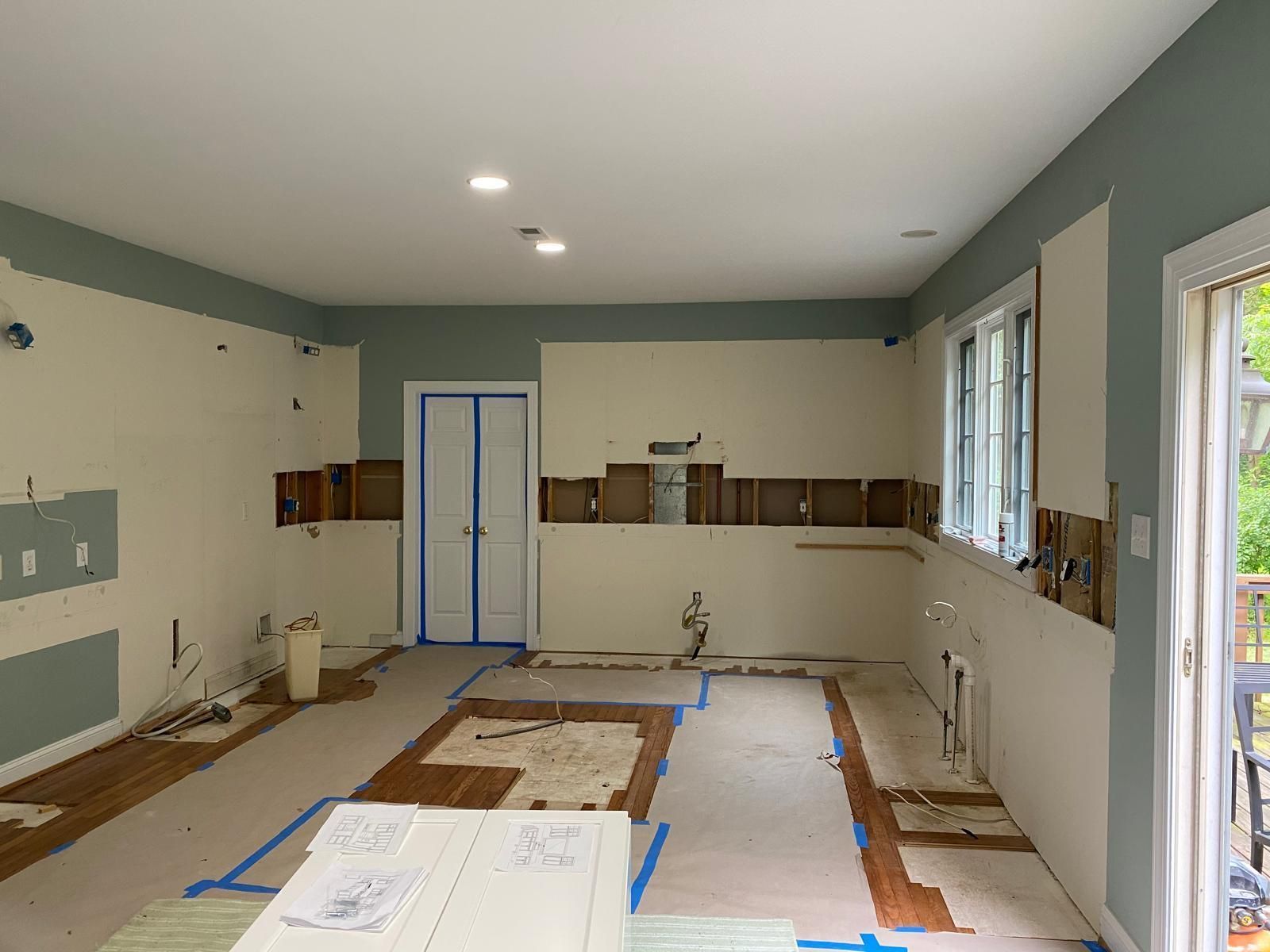 Renovation in progress in a living room with taped floor coverings, bare walls, and a windowed door.